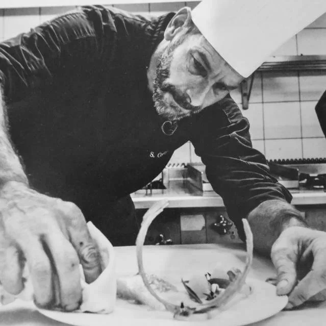 Un chef en tenue et toque blanche, concentré, décorant un plat dans une cuisine.
