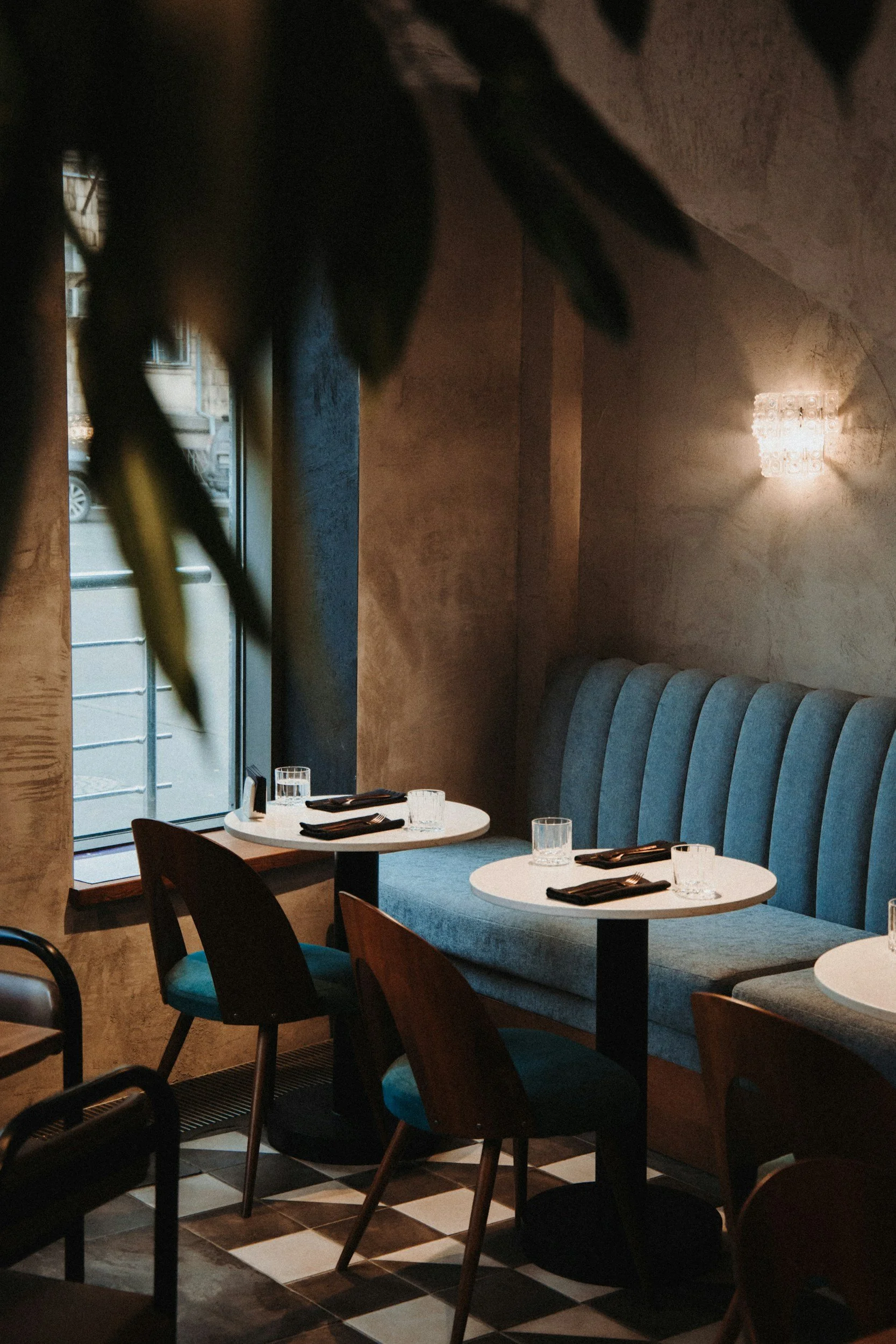 Coin salon de restaurant avec tables rondes, chaise en bois, banquette rembourrée bleue, décor moderne, lumière tamisée, vue sur la rue