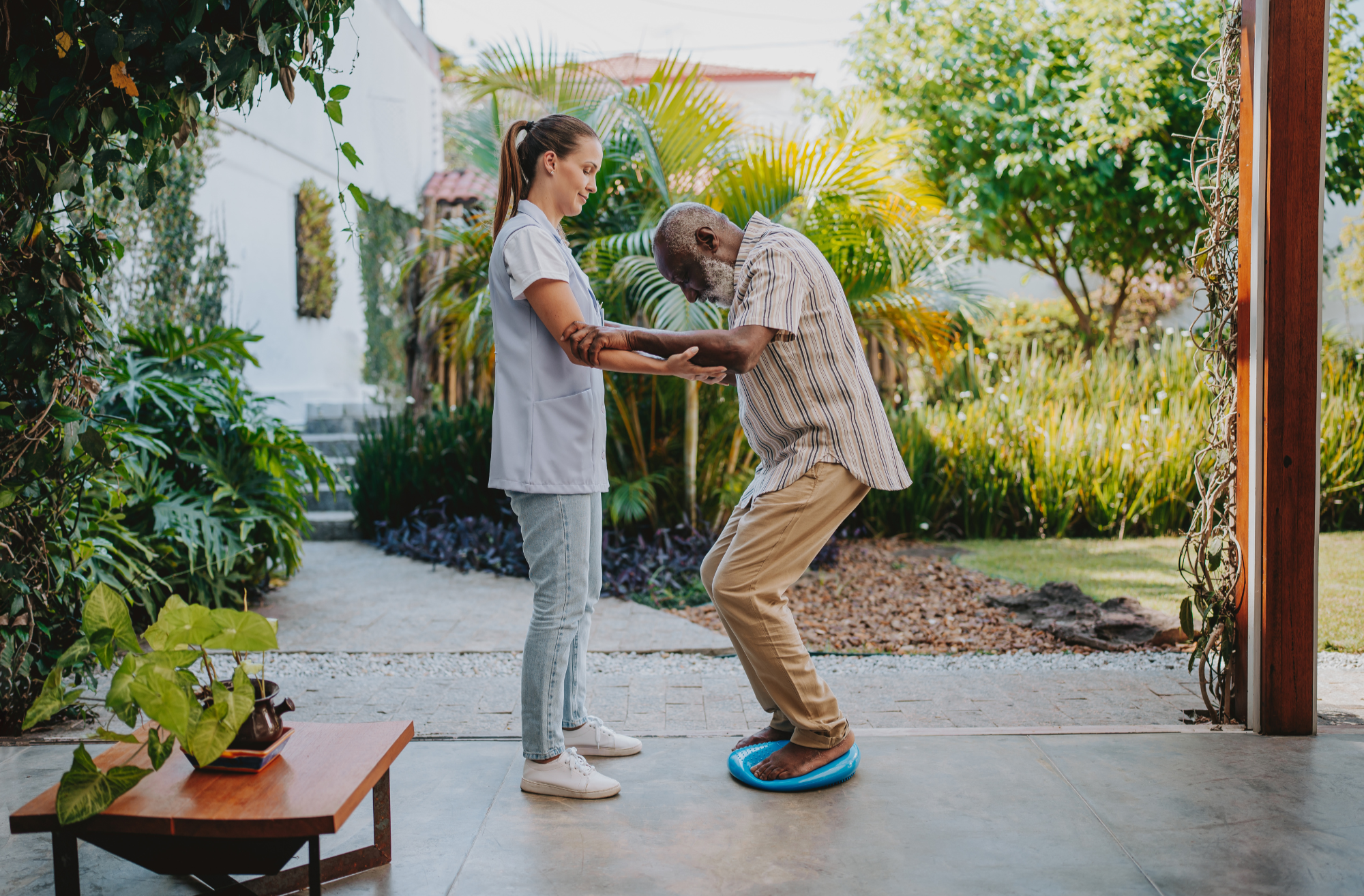 A healthcare worker, woman in scrubs, holding an elderly man's hands as he stands on a balance platform, outdoors in a garden.