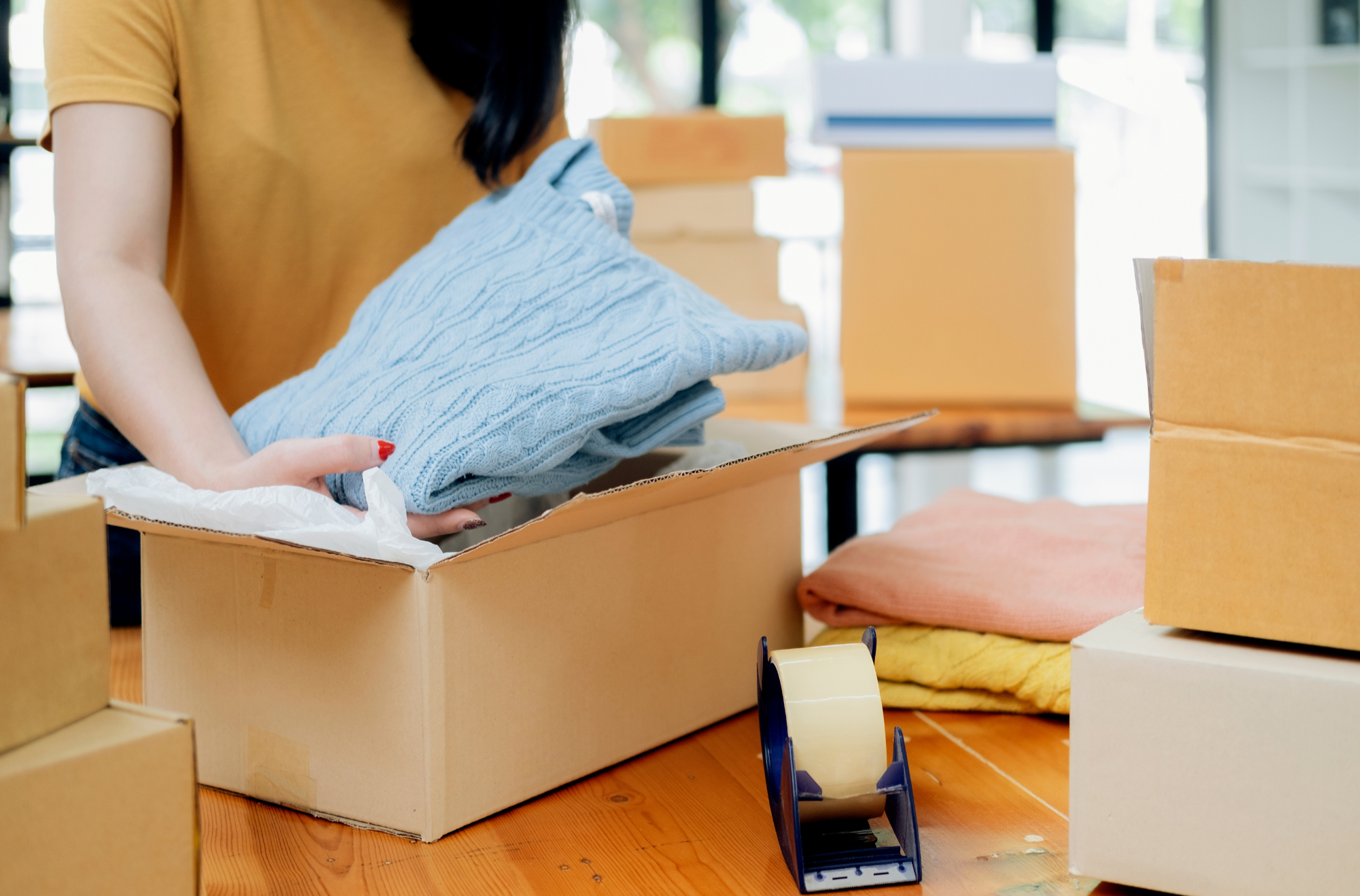 Close-up of a person in a yellow shirt packing or unpacking a box filled with folded clothes, with additional boxes and supplies around on a wooden floor.