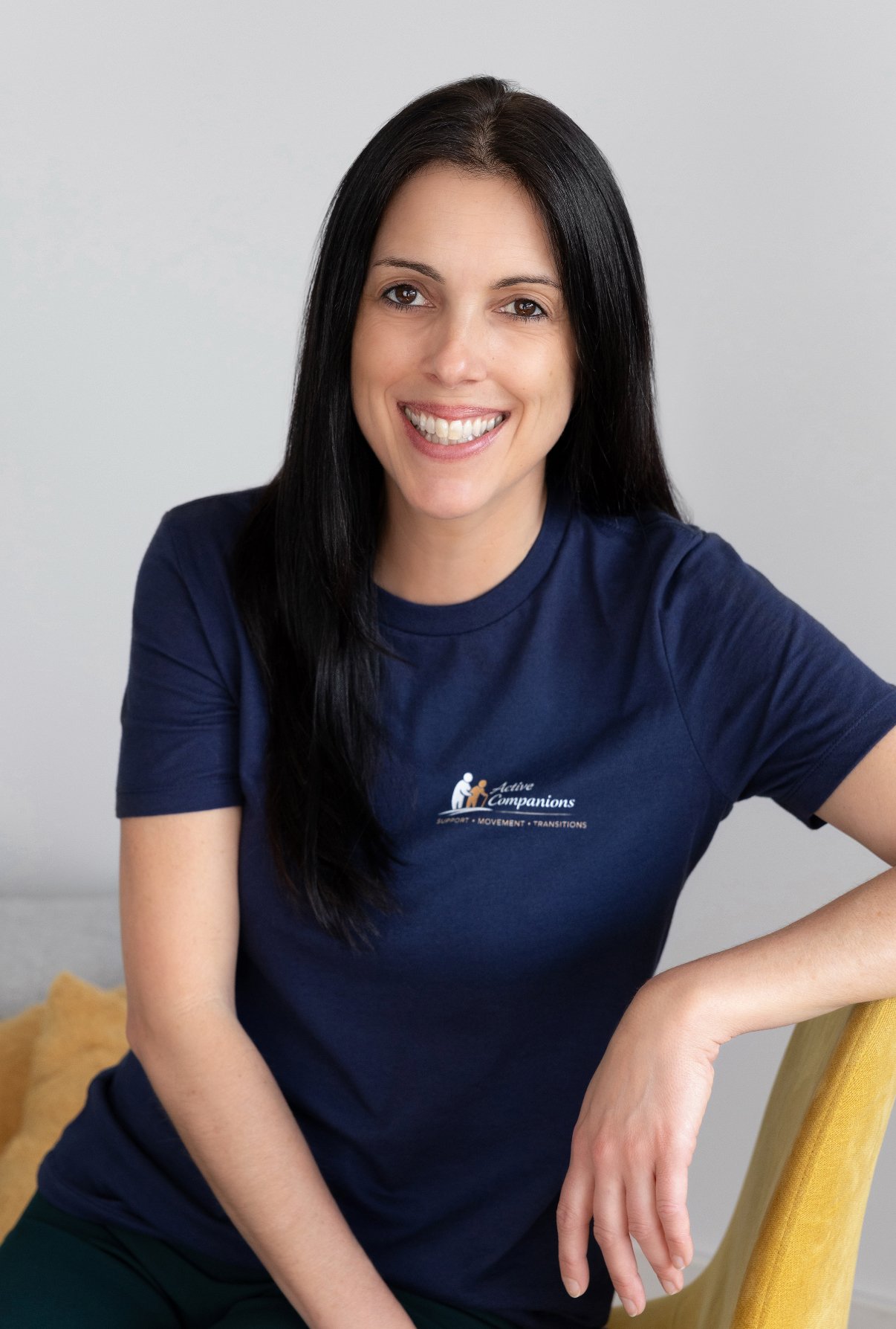 A woman with long black hair smiling, wearing a navy blue t-shirt with white text and logo, sitting on a yellow chair in front of a light gray background.
