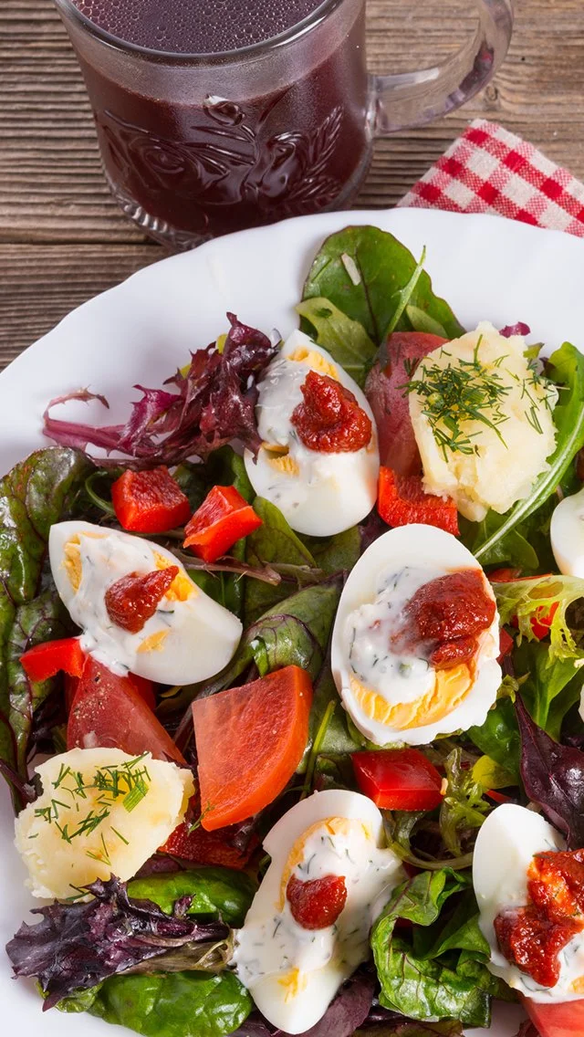 A white plate with a mixed greens salad, topped with halved boiled eggs, red bell pepper, cheese, and garnished with herbs, accompanied by a glass of dark purple juice or wine on a wooden table, with a red and white checkered napkin.
