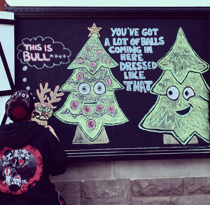 Anastasia drawing a chalk portrait of two Christmas trees. One tree is decorated & the other tree has none. The tree with no decorations says 'You've got a lot of balls coming in here dressed like that'. This was at Outer Layer Cards & Gifts.