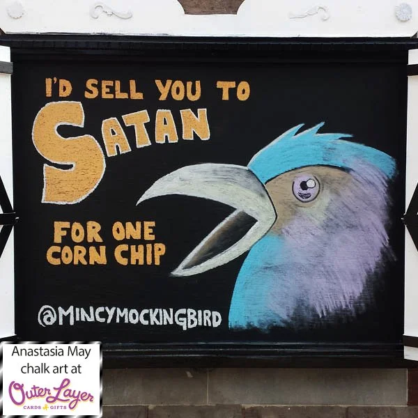 A chalk drawing of the profile of a blue, orange and pink bird screeches ‘I’d sell you to Satan for one corn chip’ and the Instagram handle of the  card brand that inspired the drawing is written under that ‘@mincymockingbird’. Anastasia May chalk ar
