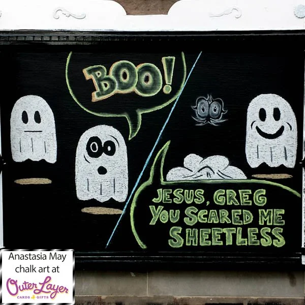 A split chalk drawing of two ghosts. The first panel shows the ghosts facing each other and one says ‘BOO’. The second panel says ‘Jesus, Greg you scared me sheetless’ and shows a sheet in a pile with floating eyes and the other ghost smiling. Anasta