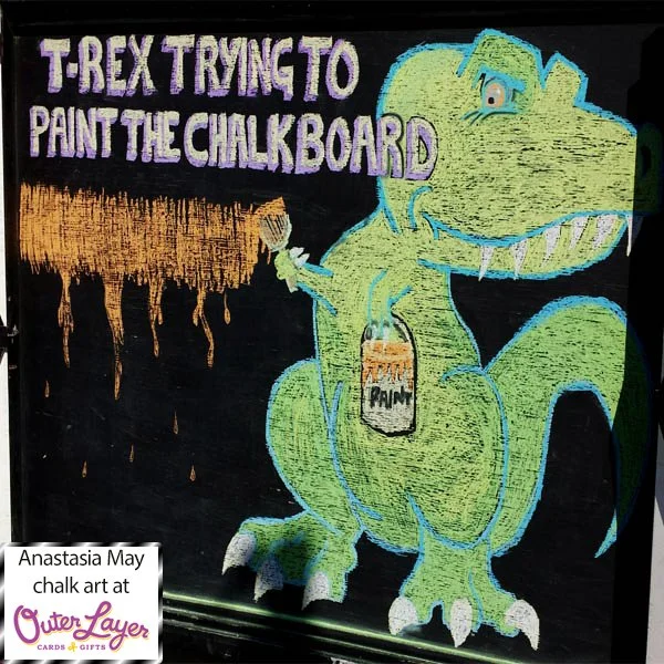 A lime green tyrannosaurus with a blue outline is drawn in chalk and holds a bucket labelled ‘paint’. The orange paint is shown in a straight line and the trex is struggling to reach further up or down with the orange paint covered brush because they