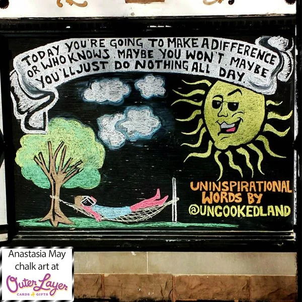 A large white banner reads ‘Today you’re going to make a difference or who knows, maybe you won’t, maybe you’ll just do nothing all day’. Under the banner there’s a chalk drawing of a sunburnt lady in a hammock asleep with a book on her face under a 