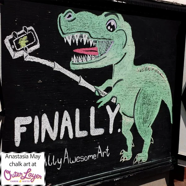 A chalk drawing of a green trex smiling holding a selfie stick to take their self portrait. The board says ‘FINALLY.’ and the Instagram handle of the card brand that inspired the drawing is written under that ‘@jollyawesomeart’. Anastasia May chalk a