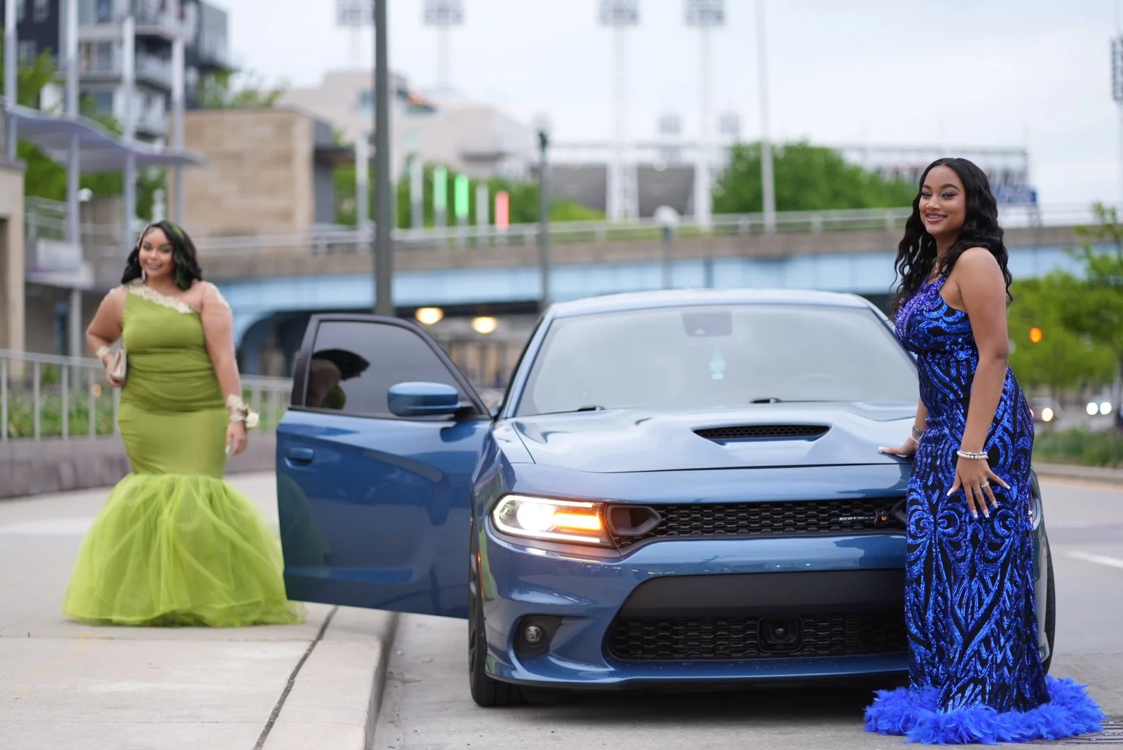 Two women in evening gowns standing near a blue sports car on a city street.