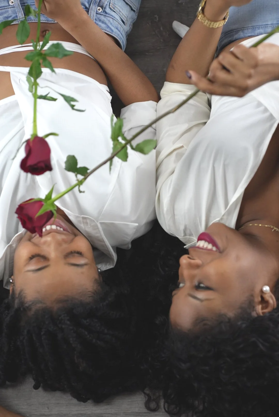 Two women lying on the floor, smiling at each other. One woman is holding a red rose, and they are dressed in white and casual clothing.