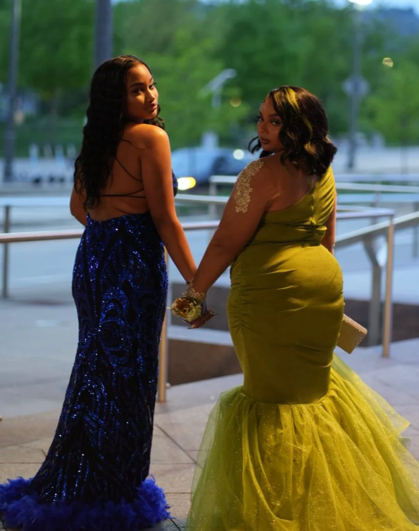 Two women dressed in glamorous evening gowns hold hands and pose outdoors during dusk. The woman on the left wears a dark blue, sequined gown with a feathered hem and backless design. The woman on the right wears a bright yellow, fitted gown with a tulle mermaid skirt and lace details on her shoulders, holding a clutch purse, with trees and street scenes in the background.