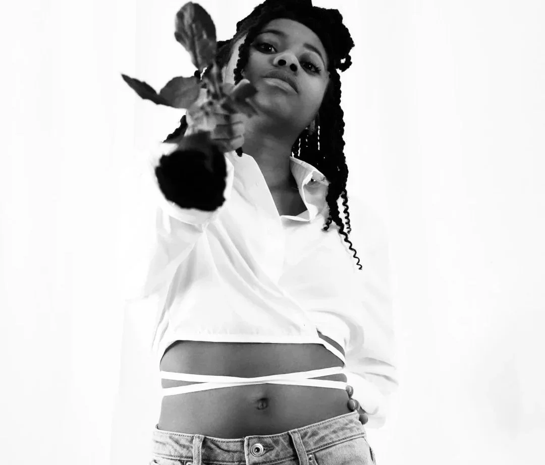 A young woman with braided hair wearing a white shirt and jeans, holding a small plant towards the camera in black and white.