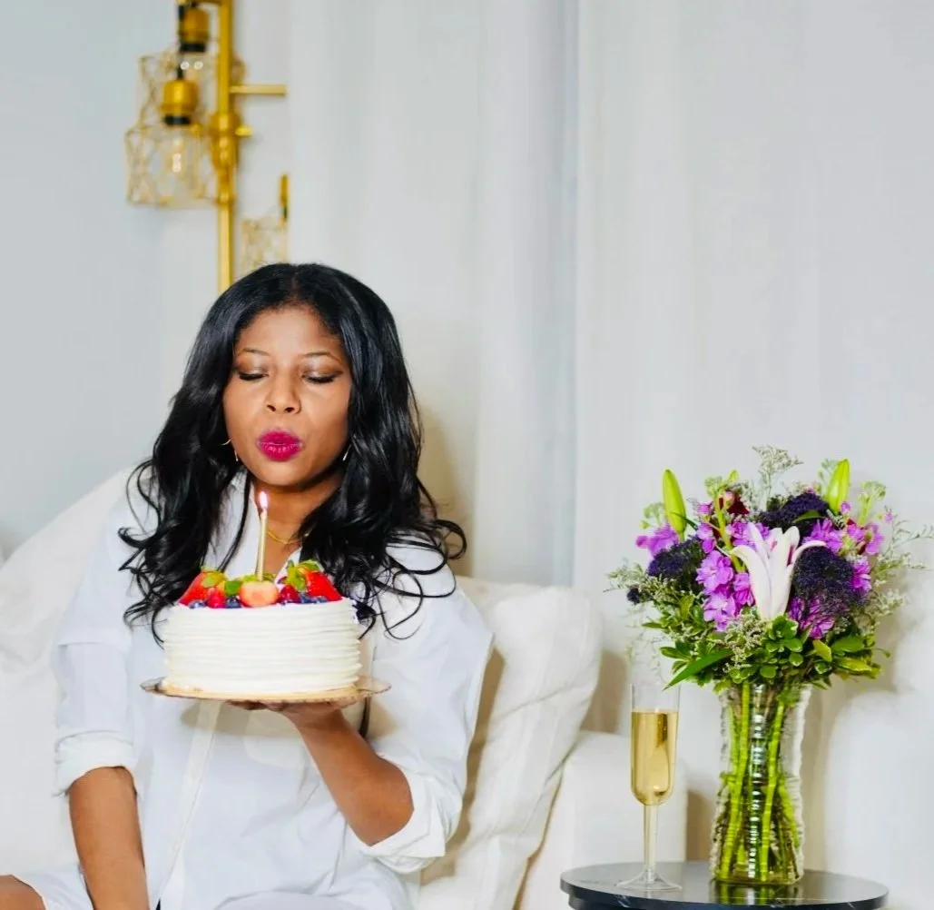 A woman with long black hair, wearing white, blowing out a candle on a birthday cake with strawberries and berries while sitting on a white sofa in a bright room with a bouquet of purple and pink flowers in a glass vase and a glass of champagne beside her.