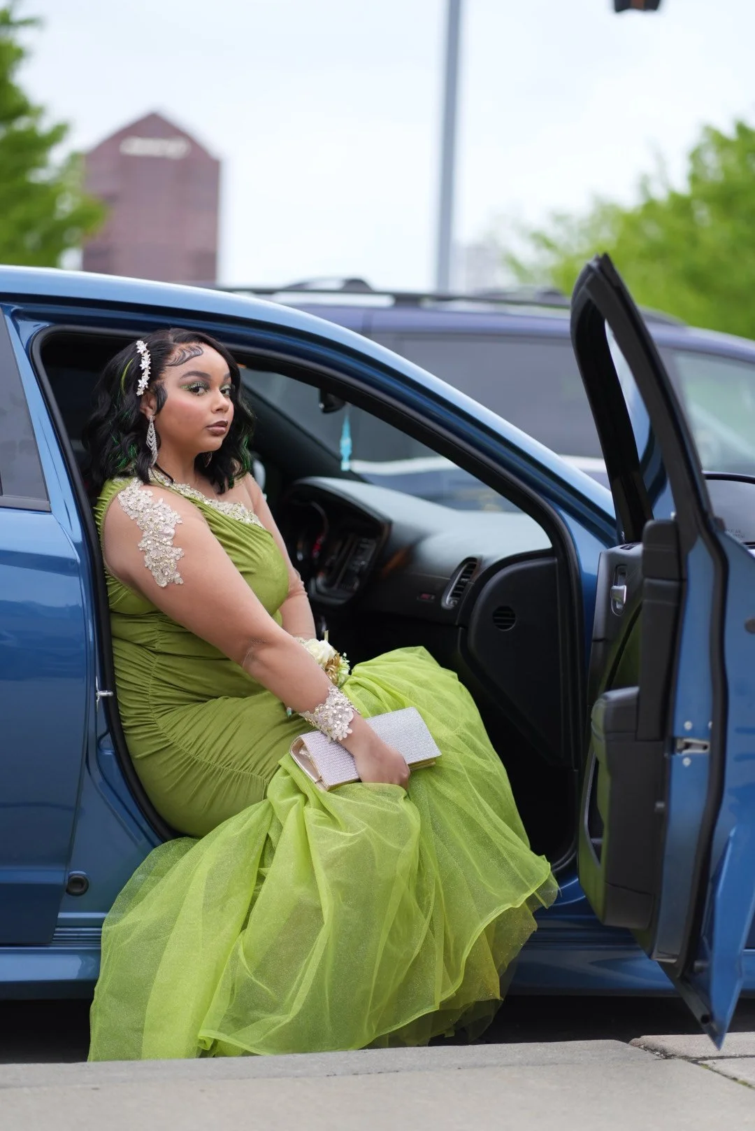 A woman in a lime green gown sitting in the passenger seat of a blue car, holding a clutch purse, with the car door open.