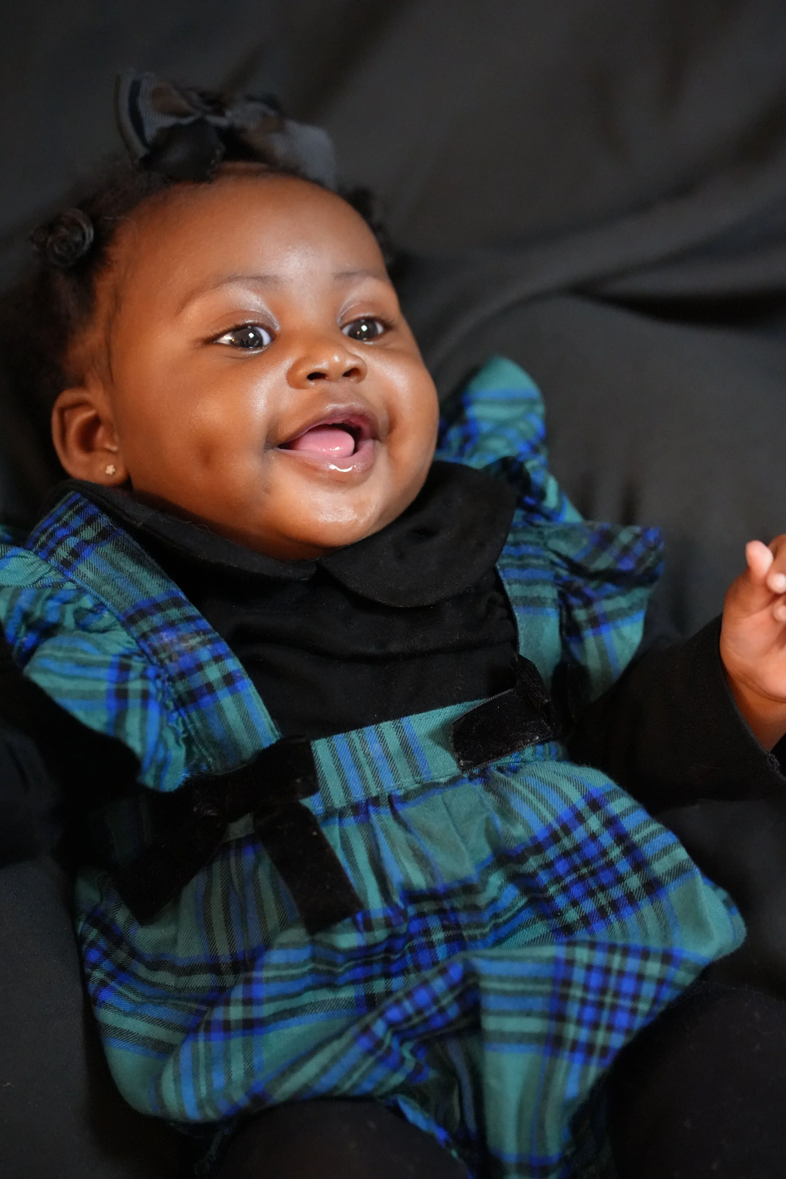 A happy African American baby girl smiling and sticking out her tongue while sitting in a car seat, wearing a blue and green plaid dress with black accents, a black collar, and a gray bow in her hair.