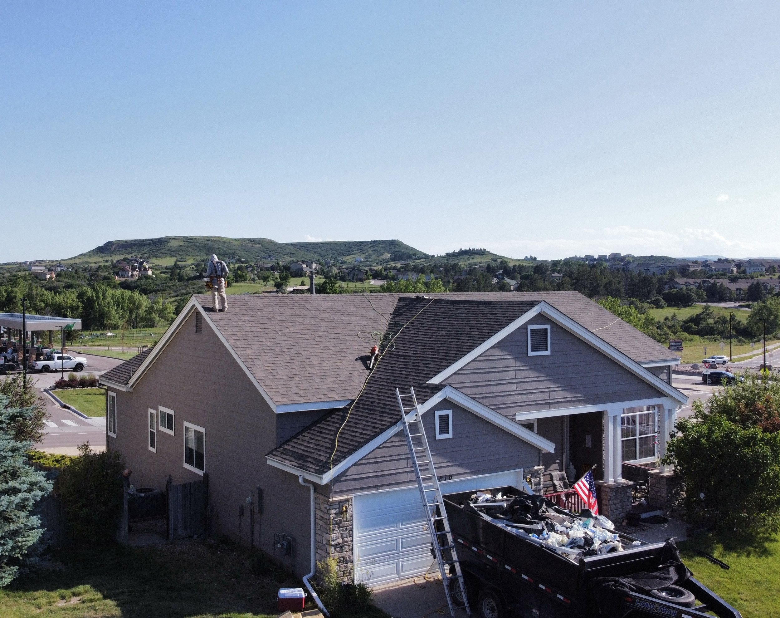 Construction workers on the roof of a house, one standing and another on a ladder, with a truck filled with debris parked in front.