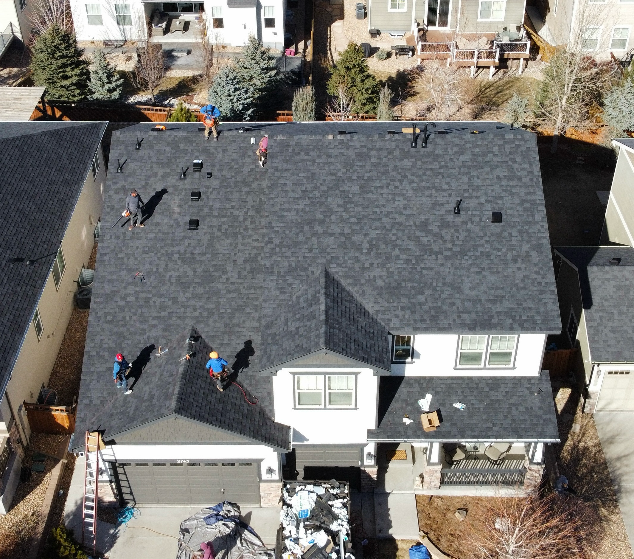 Aerial view of a house undergoing roof installation with multiple workers on the roof installing new shingles. Some workers are using safety harnesses, and the house has a gray roof with a dormer window facing the street. The driveway and front yard 