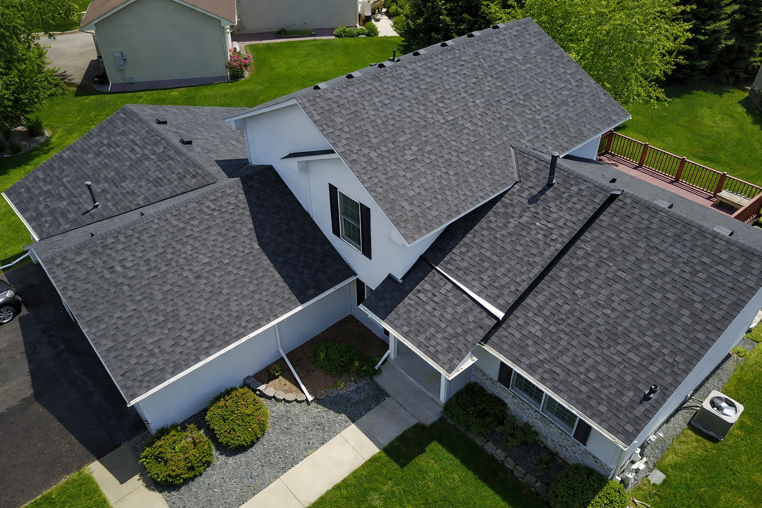 Aerial view of a residential house with multiple black-shingled roofs, a white exterior, black window shutters, a front porch with steps, a driveway with a parked vehicle, a backyard with a deck, green lawn, and trees.