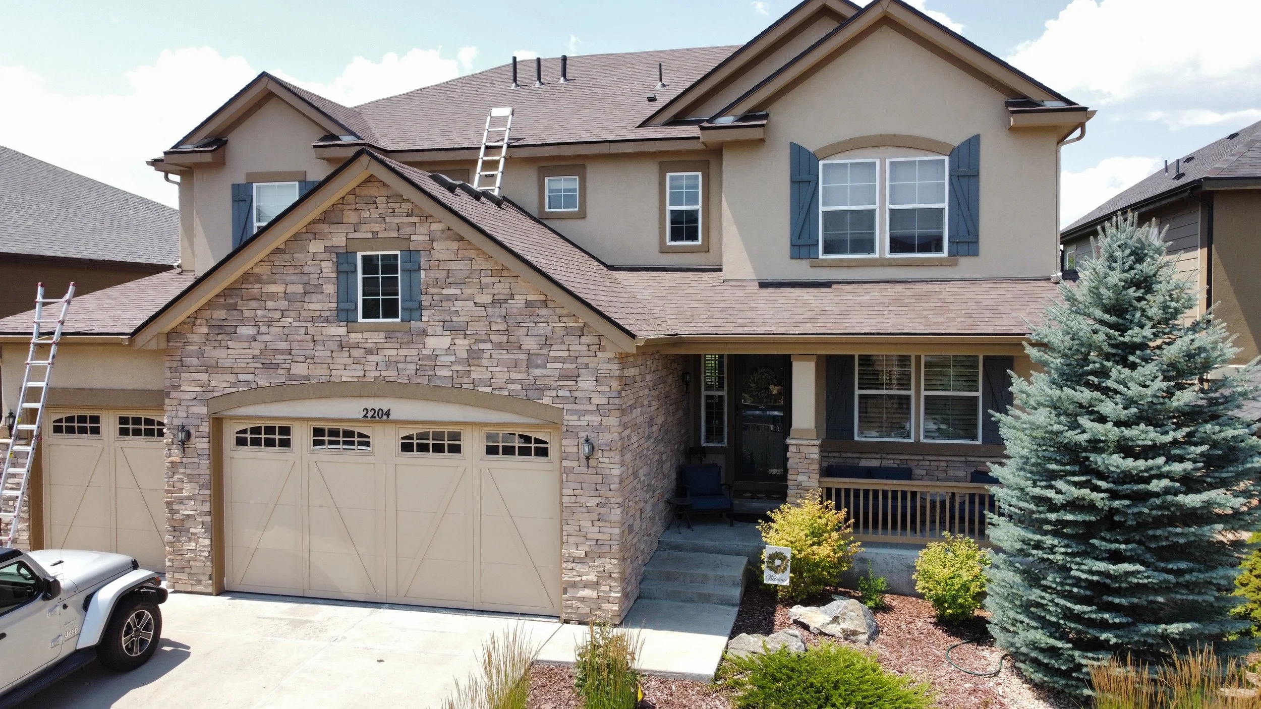 Front view of a two-story house with a stone and stucco facade, a two-car garage, a driveway, a blue evergreen tree, and a small garden with shrubs and rocks.