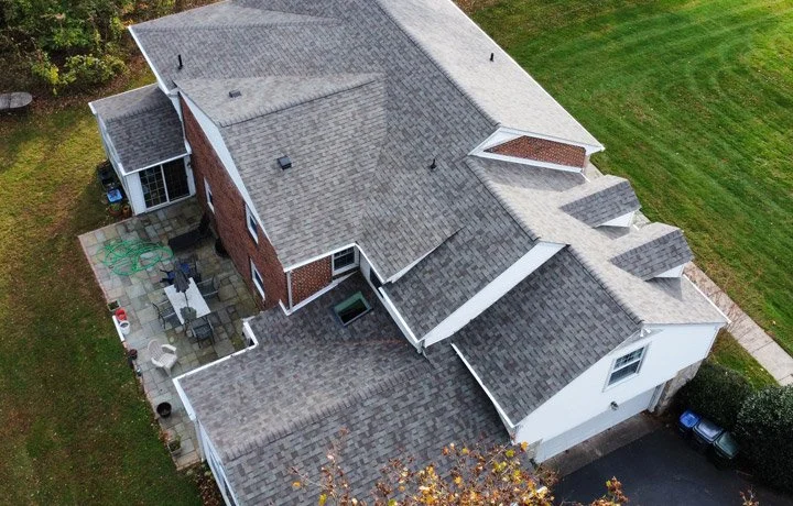 Aerial view of a large two-story house with a combination of brick and white siding, multiple roof sections, and a backyard patio with outdoor furniture and gardening hoses.