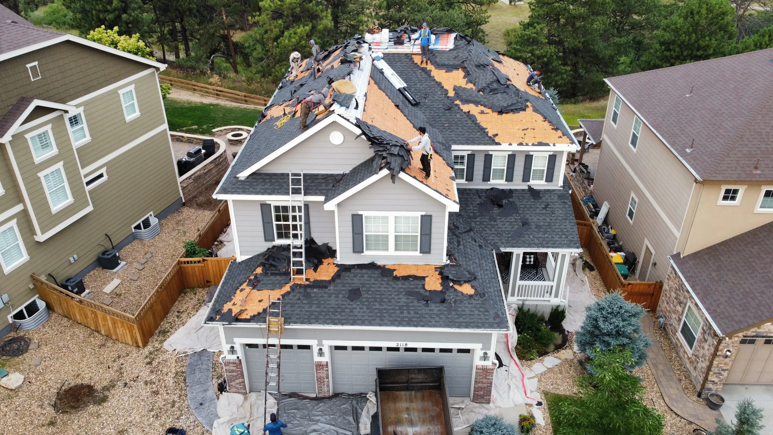 A house undergoing roof replacement with workers removing old shingles and installing new black shingles, surrounded by neighboring houses and greenery.