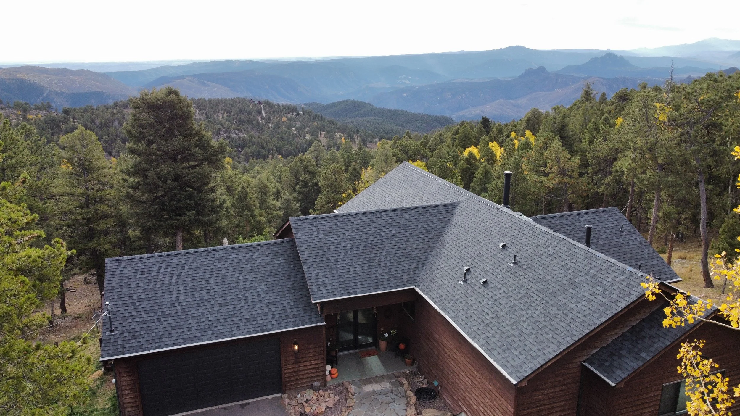 A house with a dark gray shingled roof, surrounded by a forest of tall trees, with mountain ranges in the distance under a partly cloudy sky.