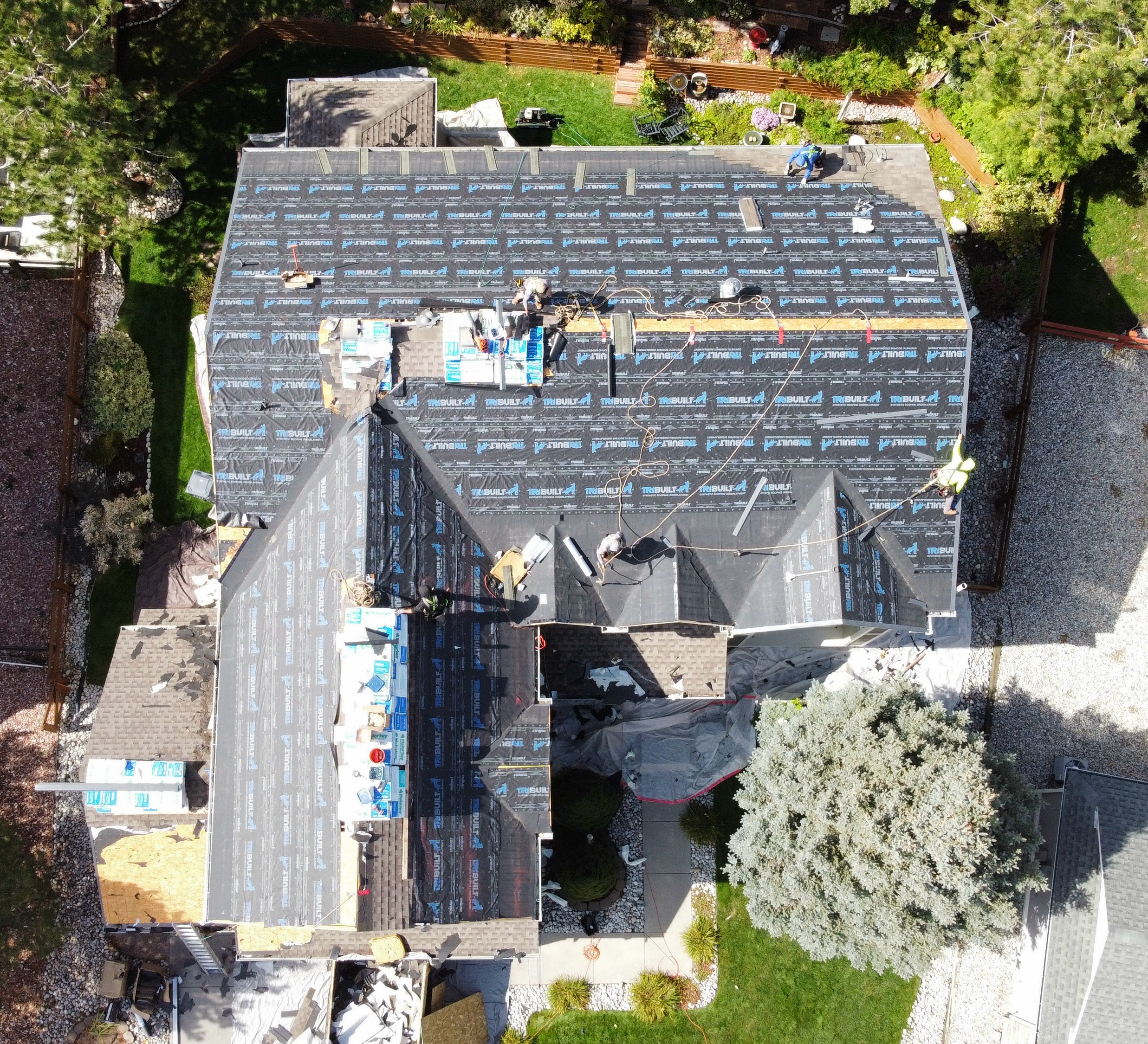 An aerial view of a house under construction with roofing workers on the roof. The roof is covered with black waterproof membrane, and tools and materials are visible. The house is surrounded by a green lawn, trees, and neighboring yards.