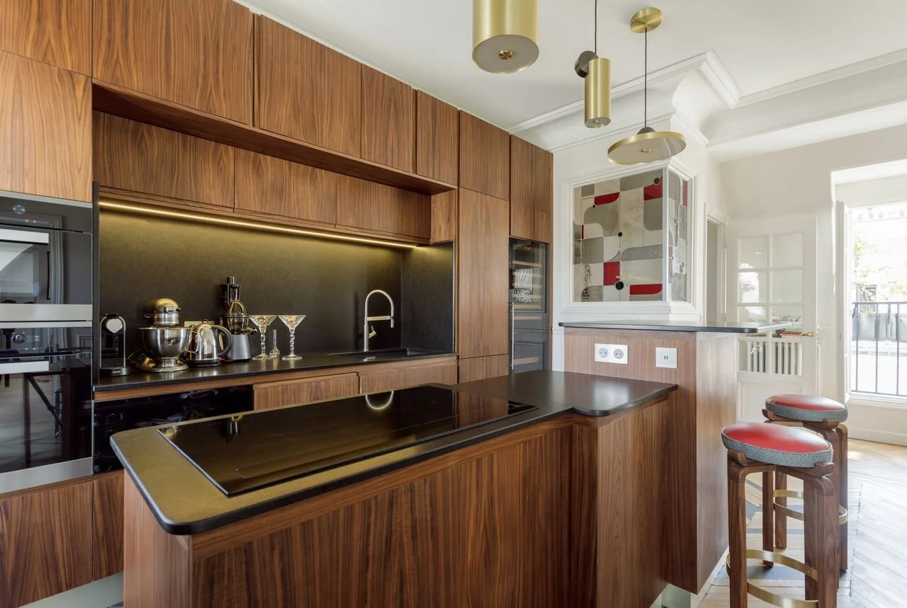 Cuisine moderne avec meubles en bois, comptoir en pierre noir et appareils en acier inoxydable, éclairage suspendu moderne, porte-fenêtre laissant entrer de la lumière naturelle.