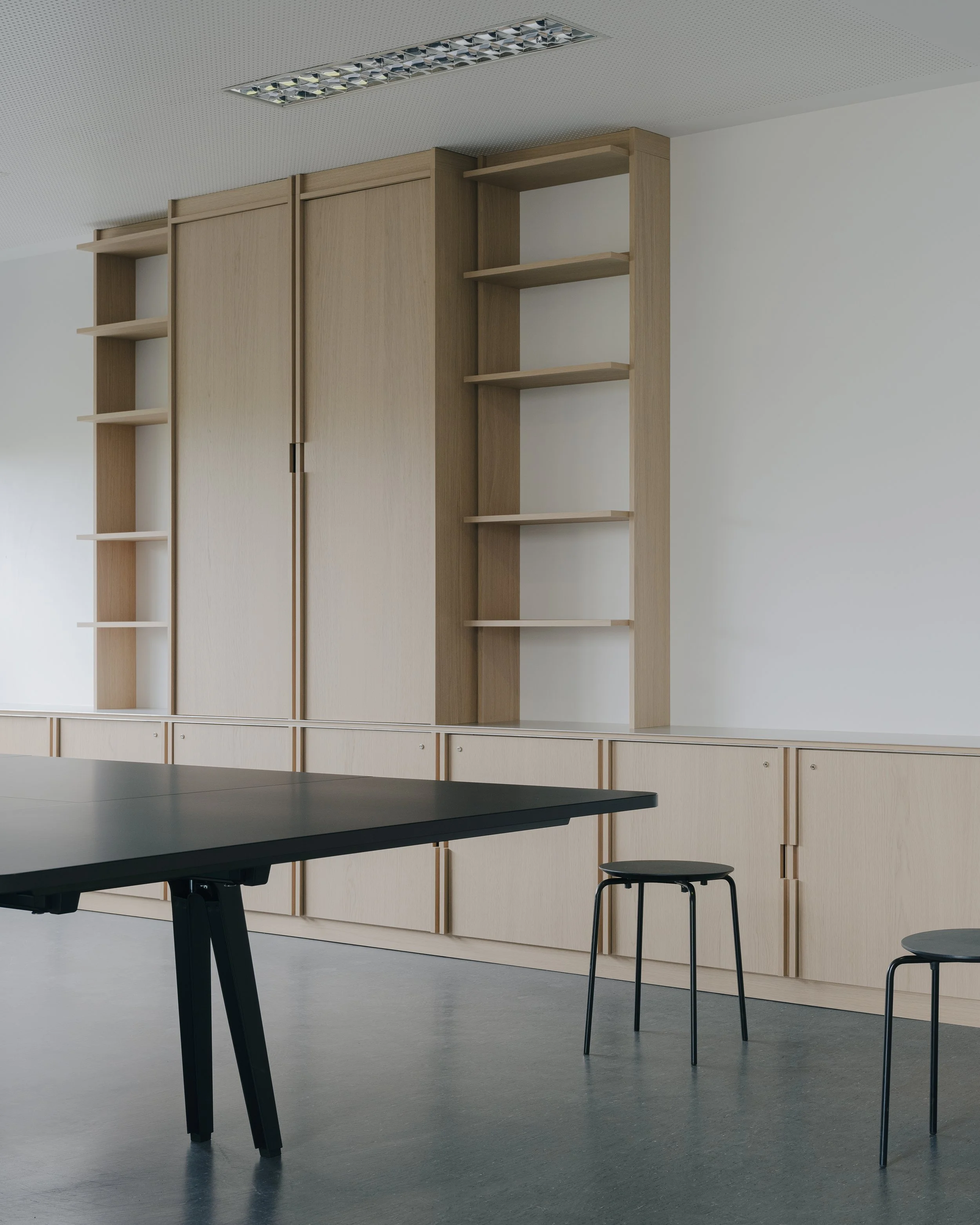 Une salle moderne avec un mobilier en bois clair, comprenant des étagères et un grand placard, ainsi que des chaises noires et une table noire