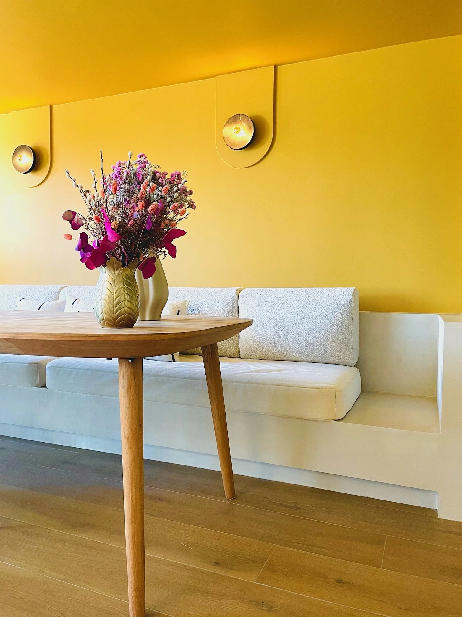 Vase avec bouquet de fleurs colorées sur une table en bois devant un canapé beige avec un mur jaune en arrière-plan