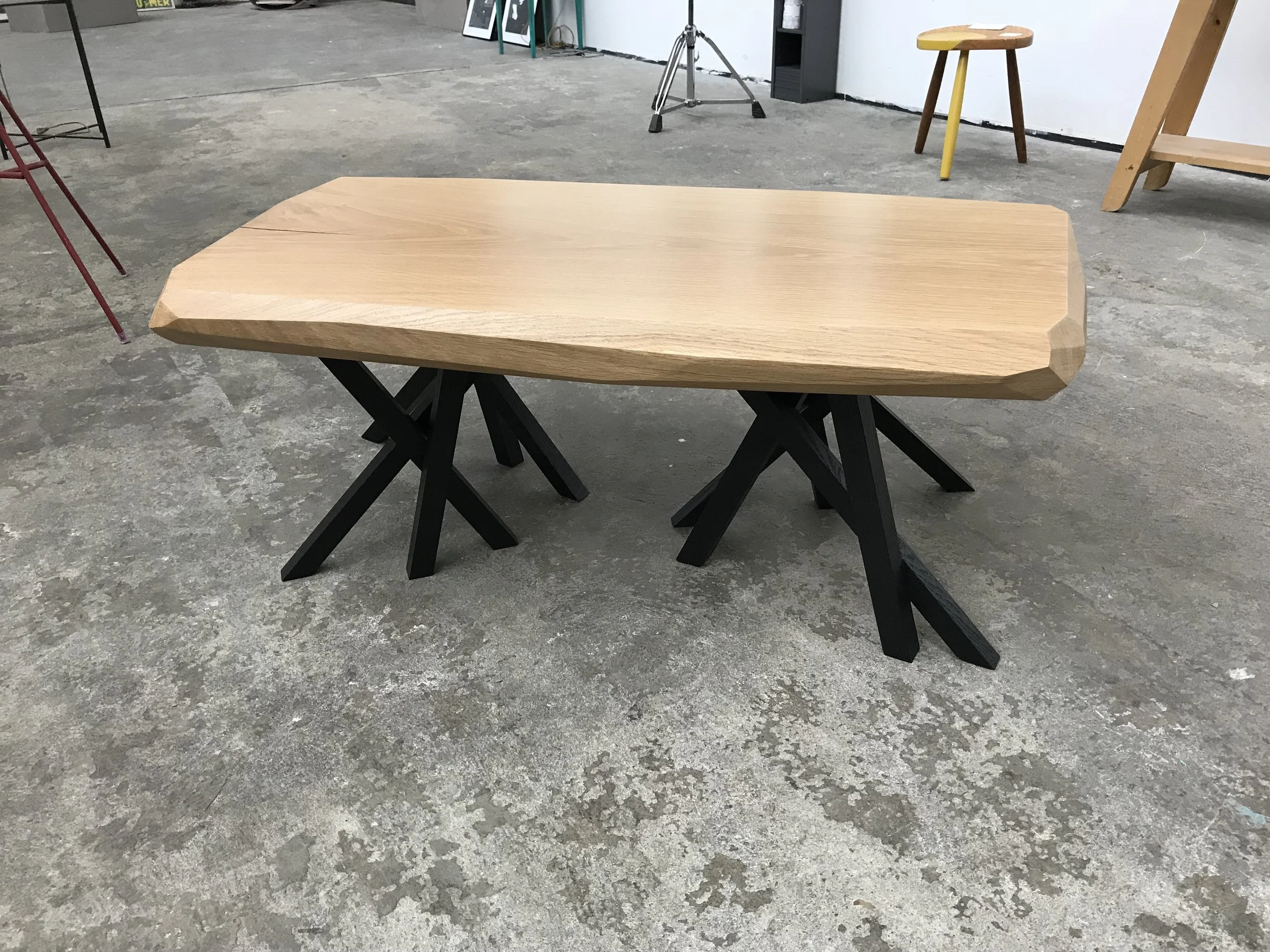 Une table en bois avec un plateau texturé et des pieds en bois noir, située dans un atelier ou un espace de travail avec un sol en béton.