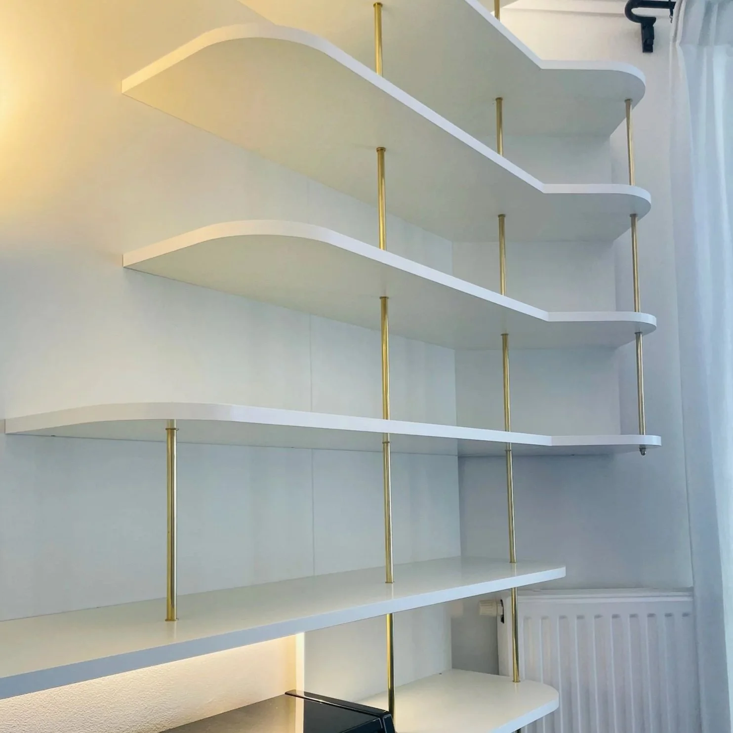 Salon avec une étagère remplie de livres, un piano noir en dessous, un fauteuil en fil métallique, une table en dessous, et des rideaux blancs fermés, décoration élégante avec moulures au plafond.