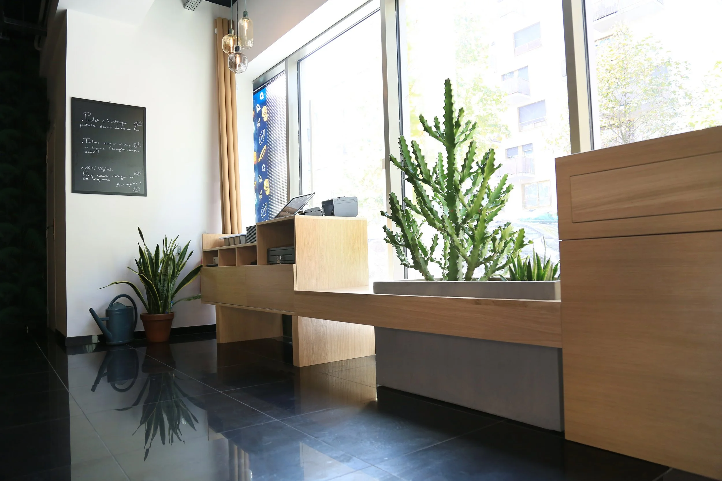 Intérieur d'un café avec des plantes vertes, une table en bois, un tableau noir avec un menu, et de grandes fenêtres laissant entrer la lumière.