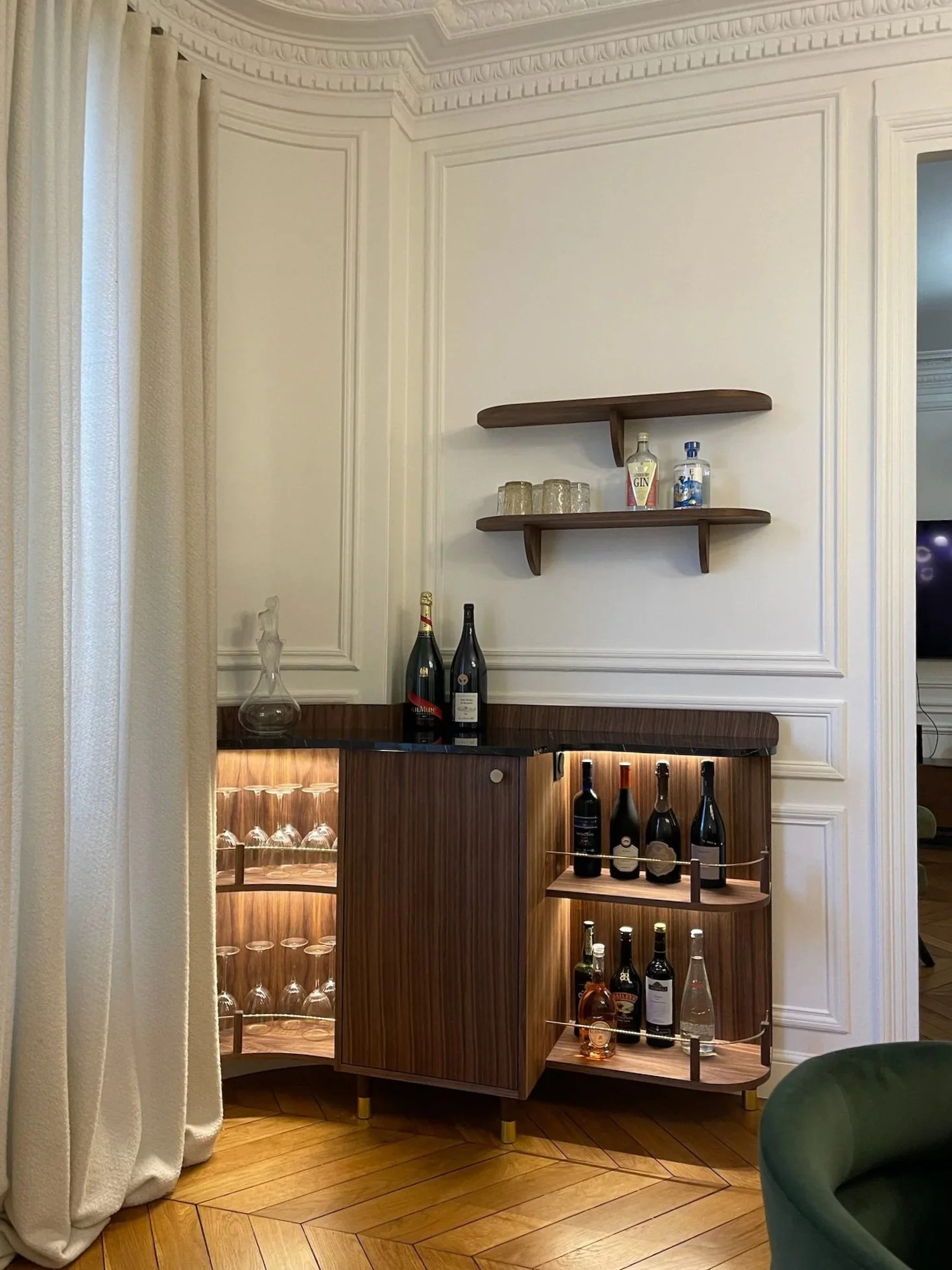 Un bar en bois sombre avec des étagères éclairées, des bouteilles de vin, des verres et un décor élégant dans une pièce aux murs blancs avec moulures et un sol en bois chaleureux.