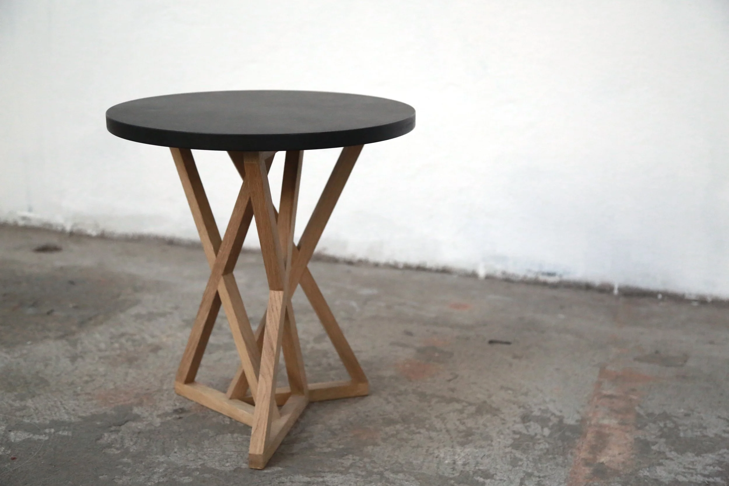 Table basse avec un plateau rond noir et une structure en bois entrecroisée, posé sur un sol en ciment dans une pièce vide.