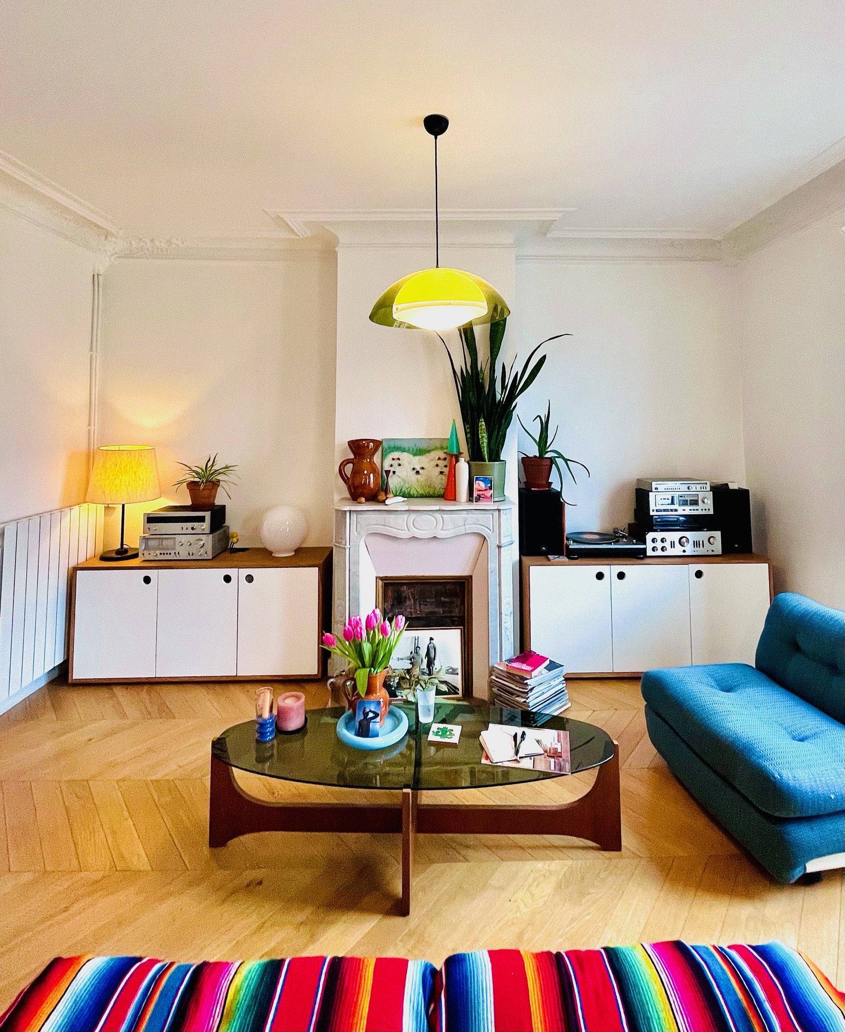 Salon avec canapé bleu, table basse en bois foncé, cheminée décorative, plantes vertes, et une station de musique. Le sol est en parquet, et un tapis coloré en rayures est visible au premier plan.