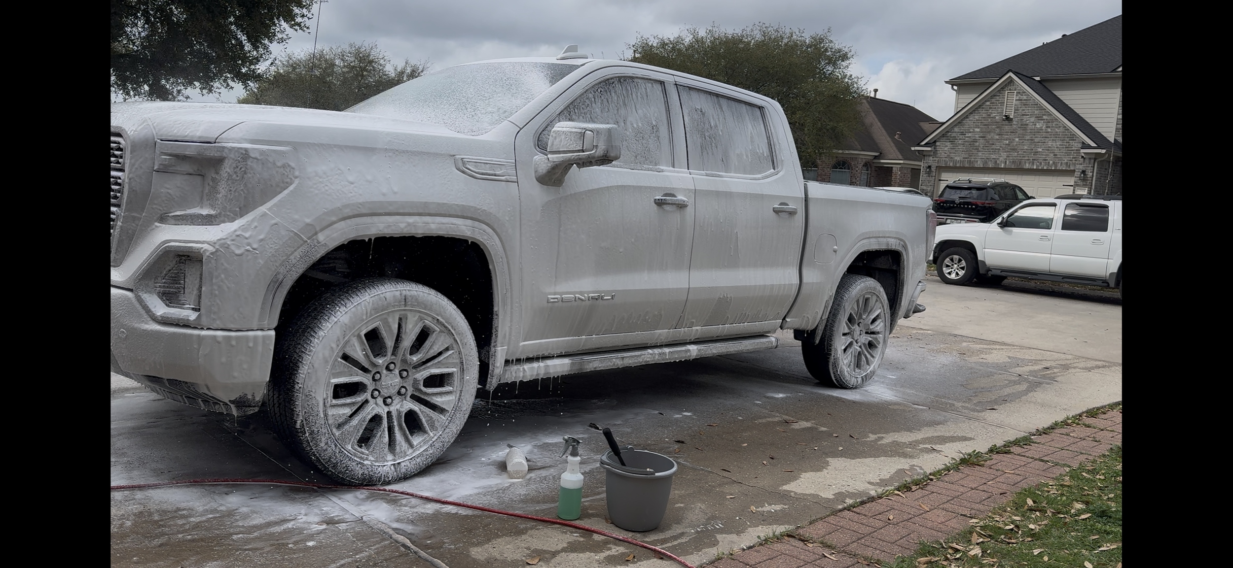 A white pickup truck is covered in soap suds and foam while being washed outside on a driveway, with cleaning supplies and a bucket on the ground nearby.