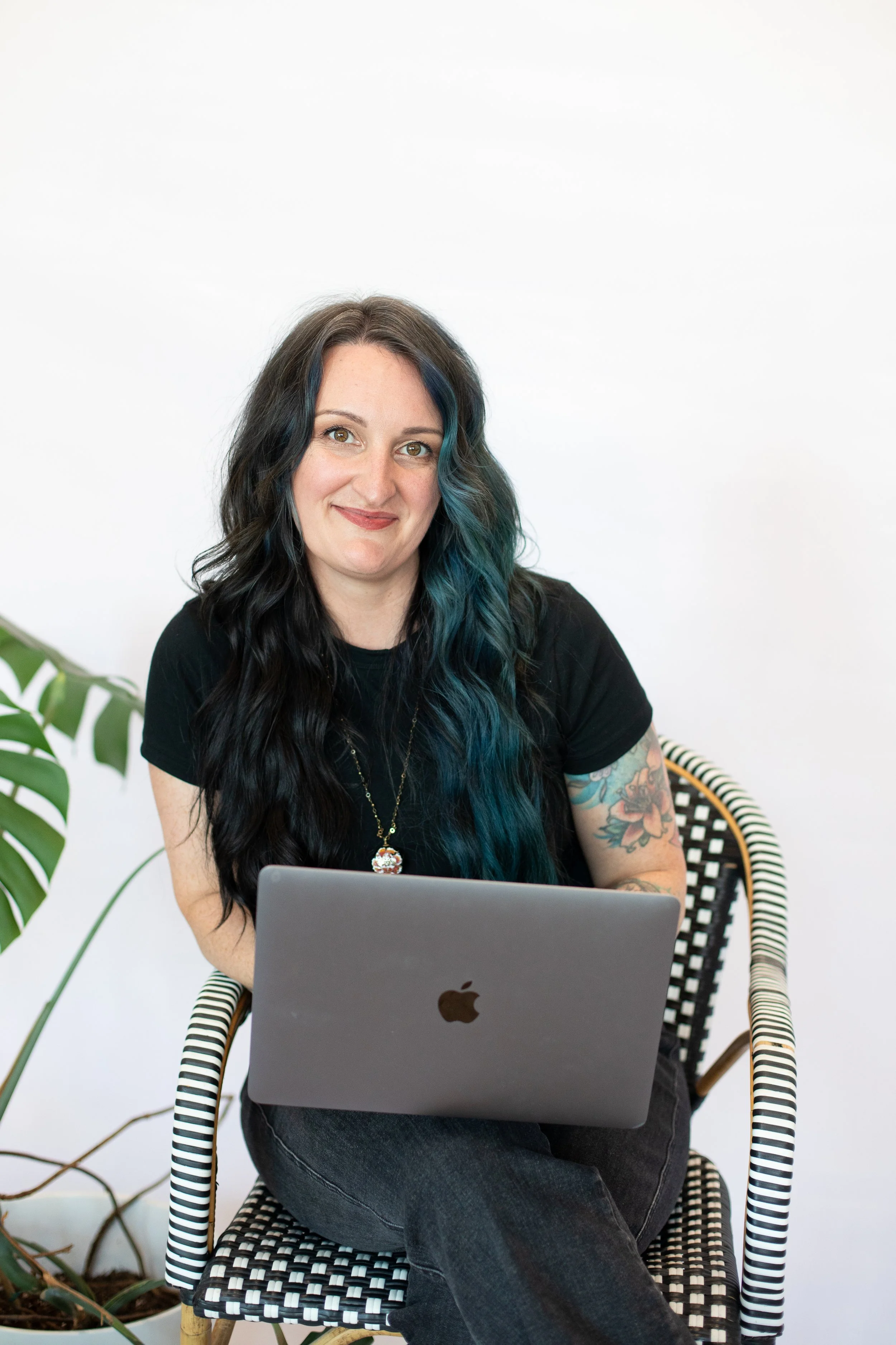 A woman with long dark hair with blue streaks, smiling, sitting in a black and white patterned chair in front of a laptop, with a large green plant to her left and a white wall background.