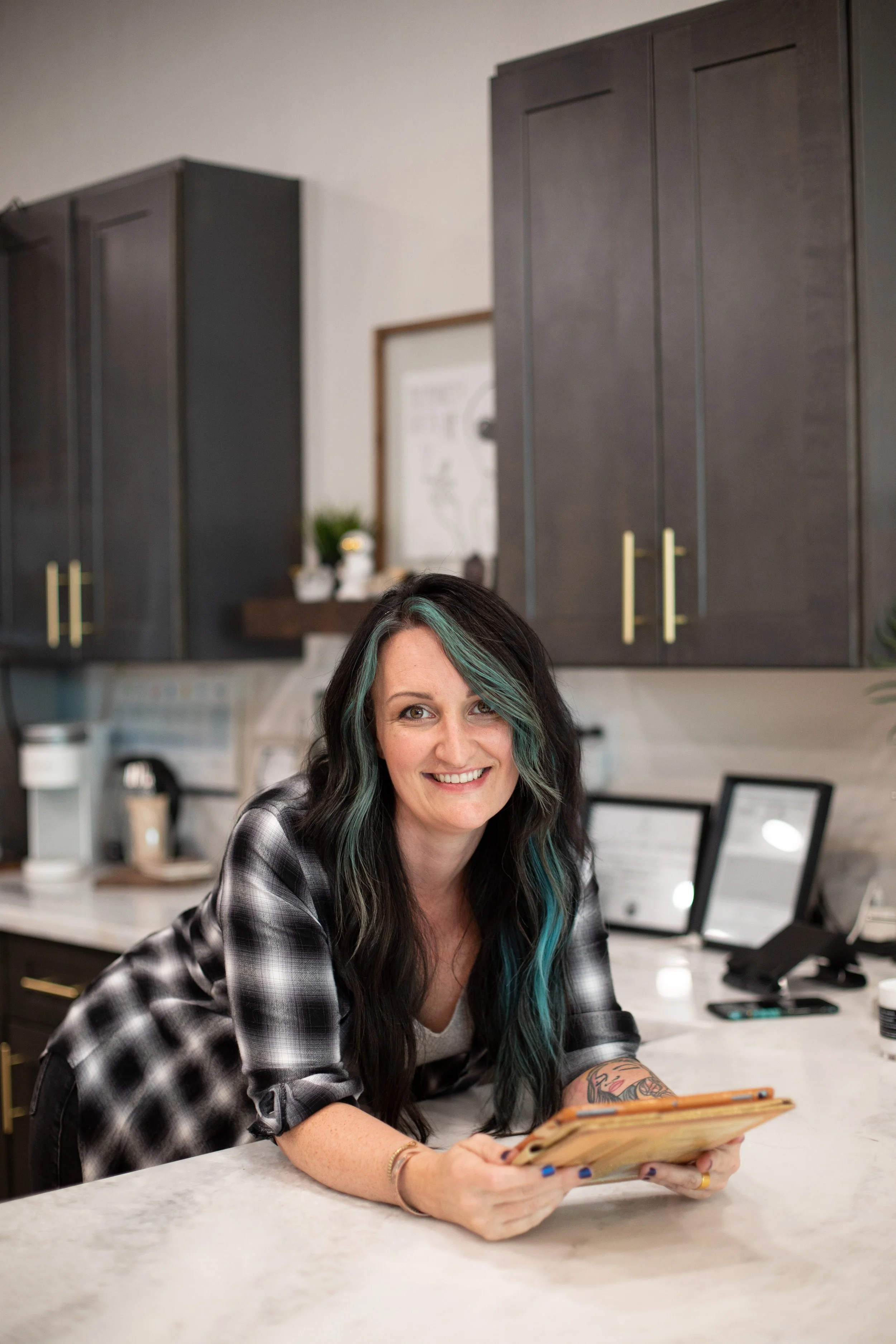A woman with black hair with teal highlights, smiling, sitting at a white marble countertop, wearing a black and white plaid shirt, holding a menu, in a modern kitchen. There's a tattooed arm visible, with gold jewelry on one wrist. Background shows black cabinets, framed pictures, a small plant, and a framed diagram.