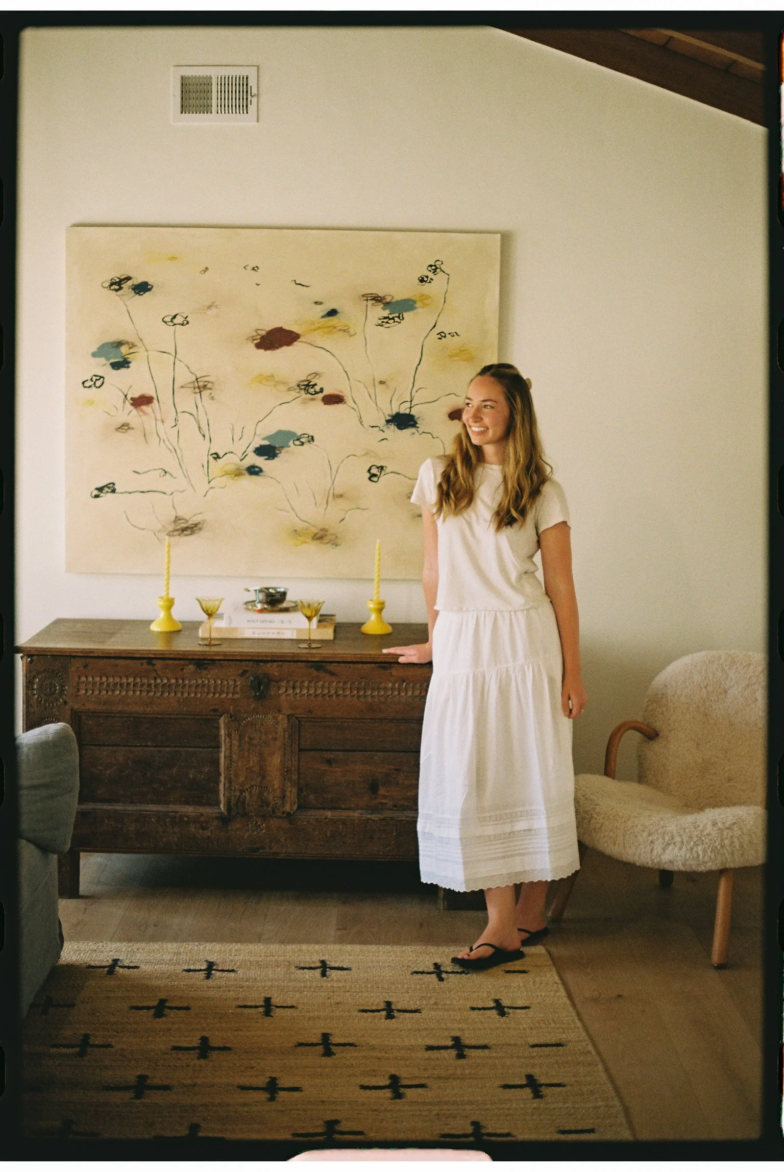 A young woman with long blonde hair wearing a white dress and black sandals standing in a living room, smiling and looking to her left. Behind her is a wall with abstract floral artwork, a wooden sideboard with candles, cups, and books, and a cozy beige armchair.