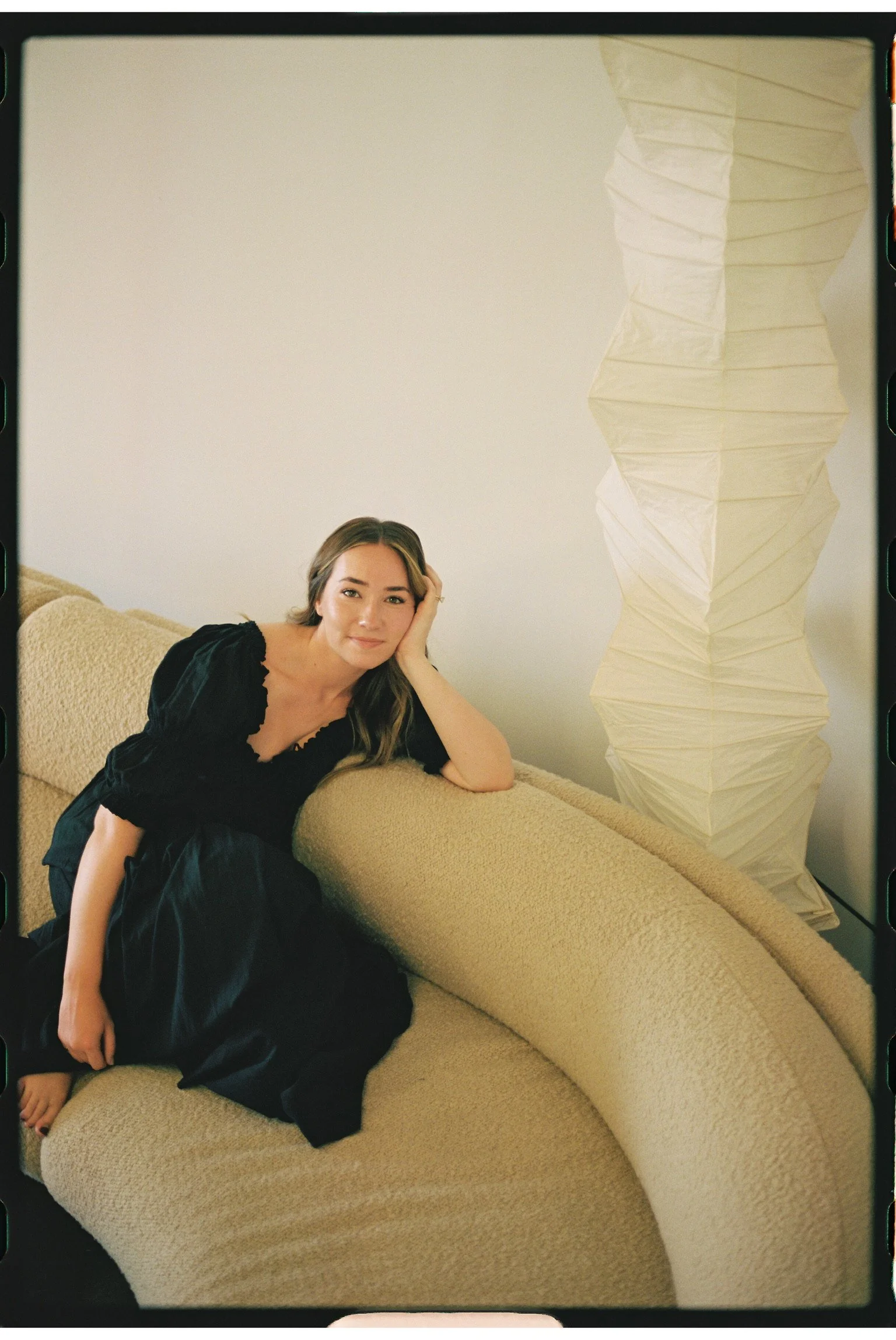 A young woman with light brown hair, wearing a black dress, is sitting on a beige, curved sofa. She rests her head on her left hand and smiles softly at the camera. There is a tall, white, crumpled-paper lamp beside her in a minimalistic room with plain white walls.
