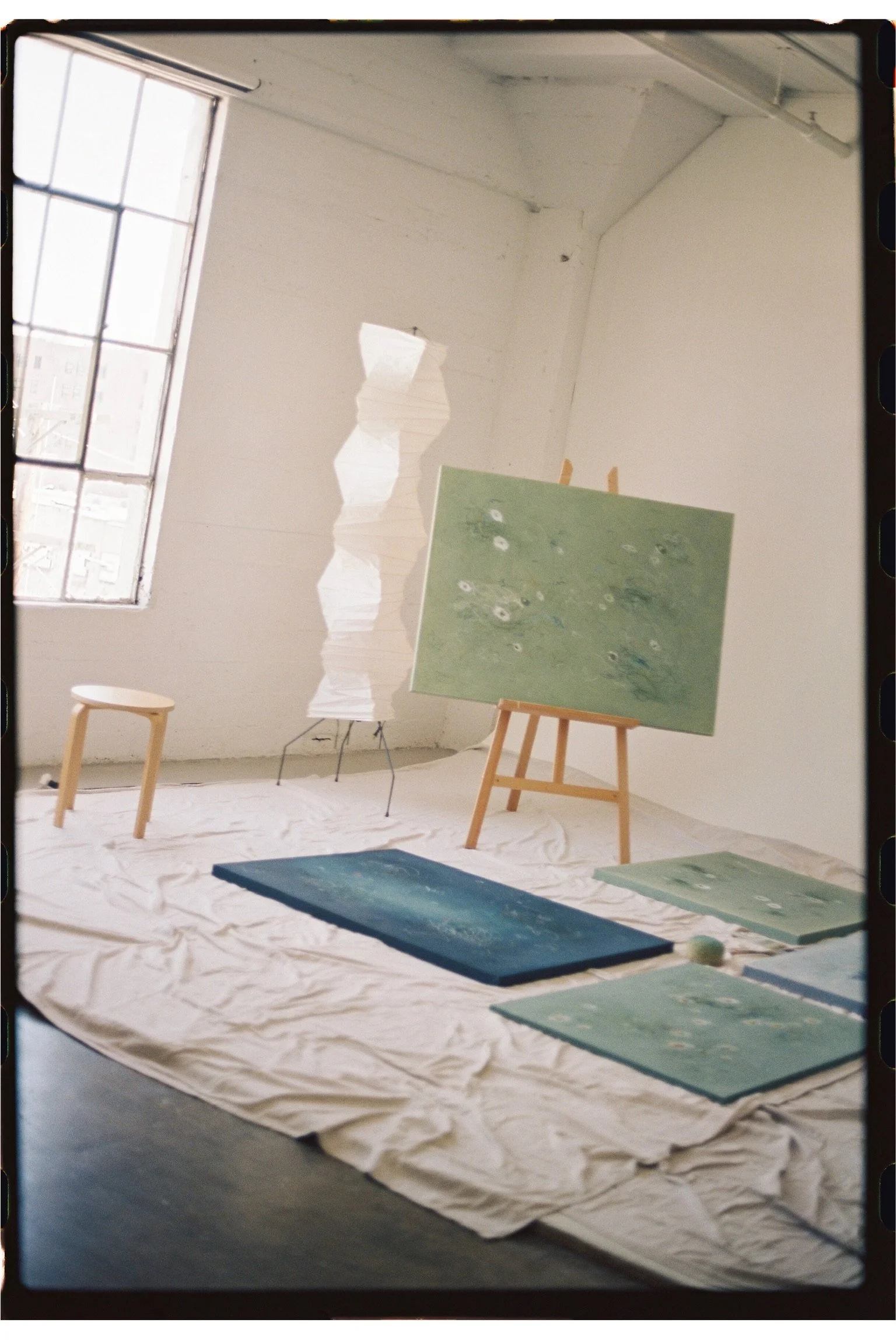 An art studio with multiple paintings of abstract, ocean-inspired themes, placed on an easel and spread around on the floor. A stool and a folded paper lantern are visible near a large window, letting in natural light.