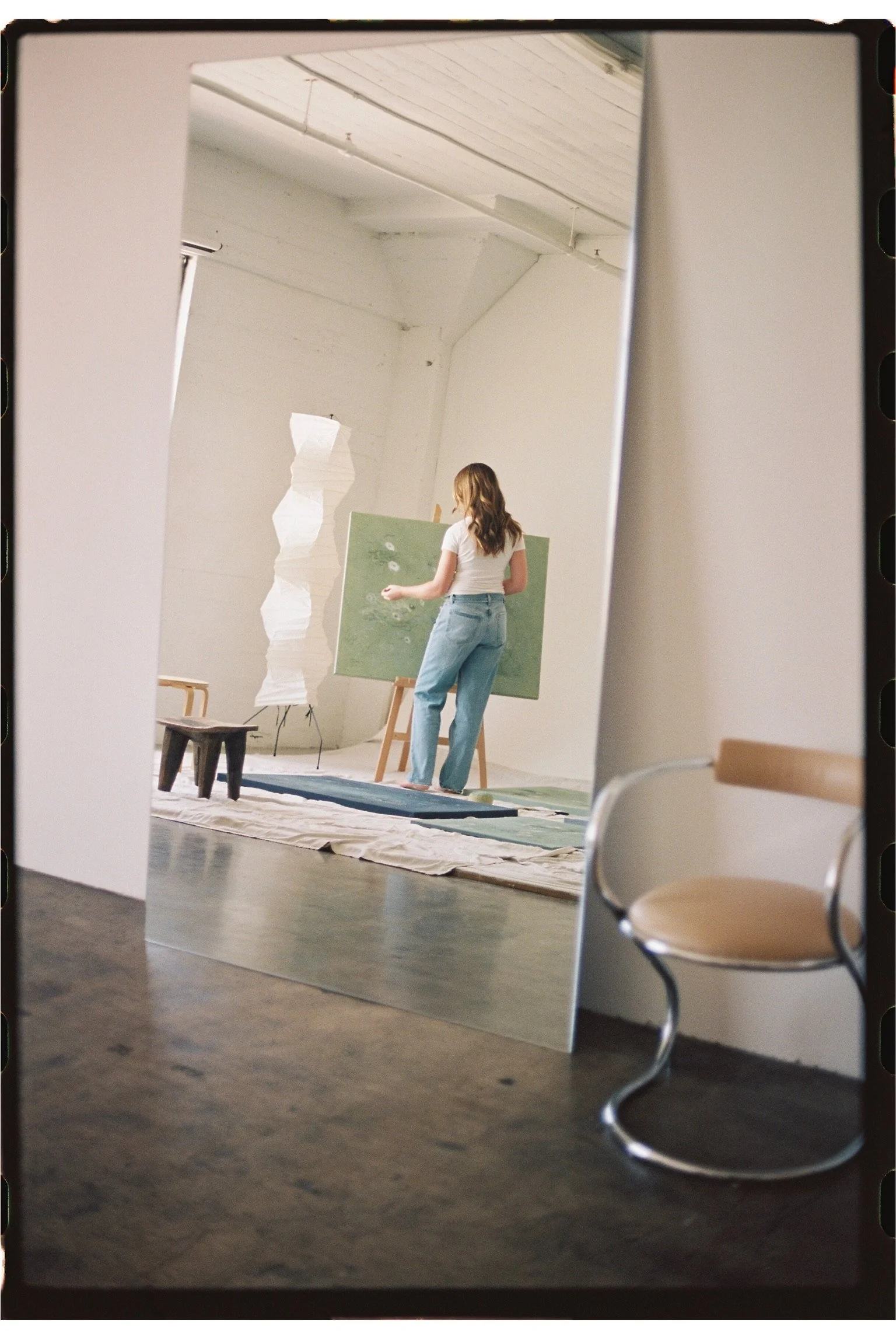 A woman standing in an art studio in front of an easel painting a green canvas, with a white textured floor lamp and a small wooden stool nearby, reflected in a large mirror leaning against a white wall.