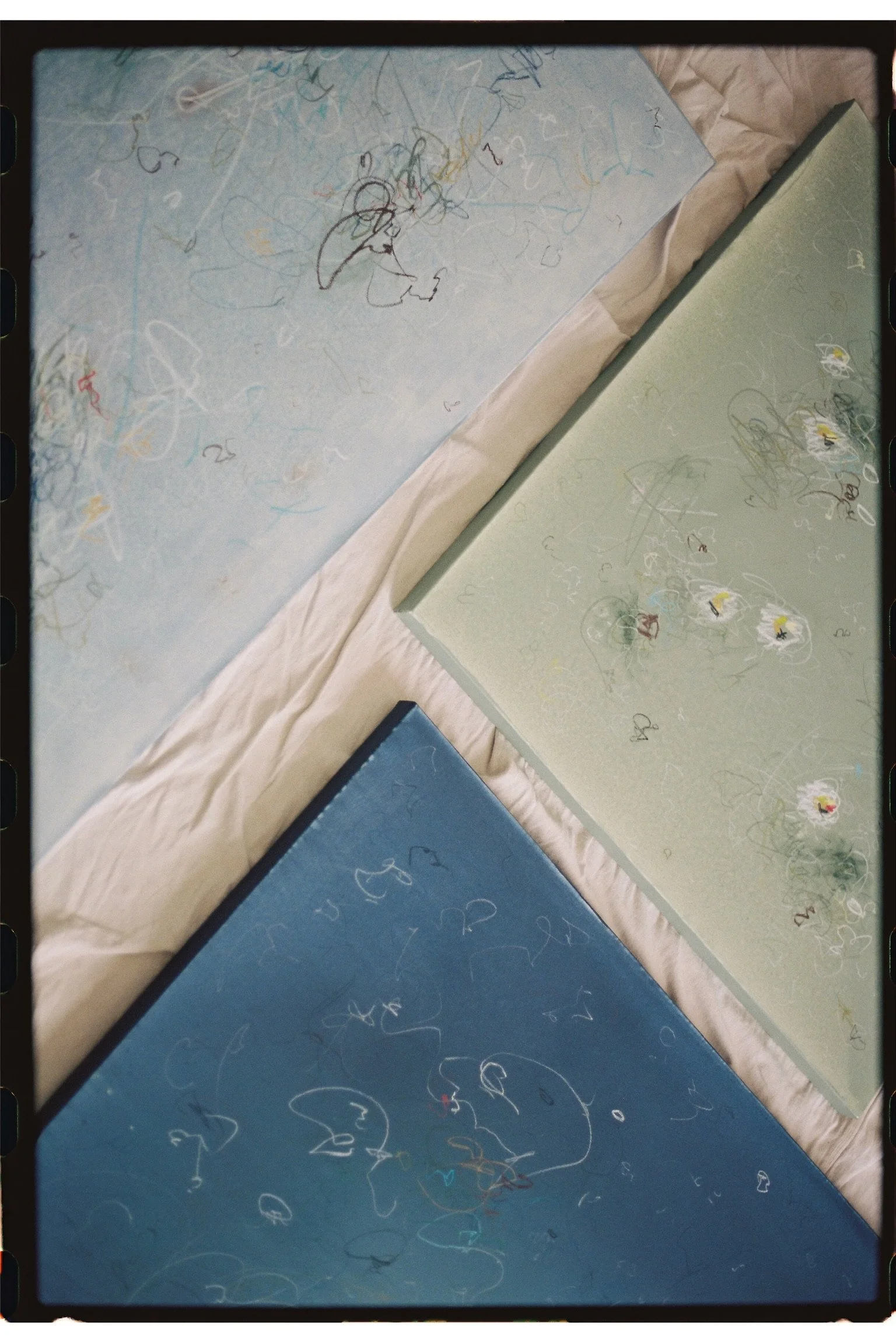 Three pastel-colored cardboard panels (blue, green, and light blue) with scribbled lines and doodles, spread out on a light-colored fabric surface.