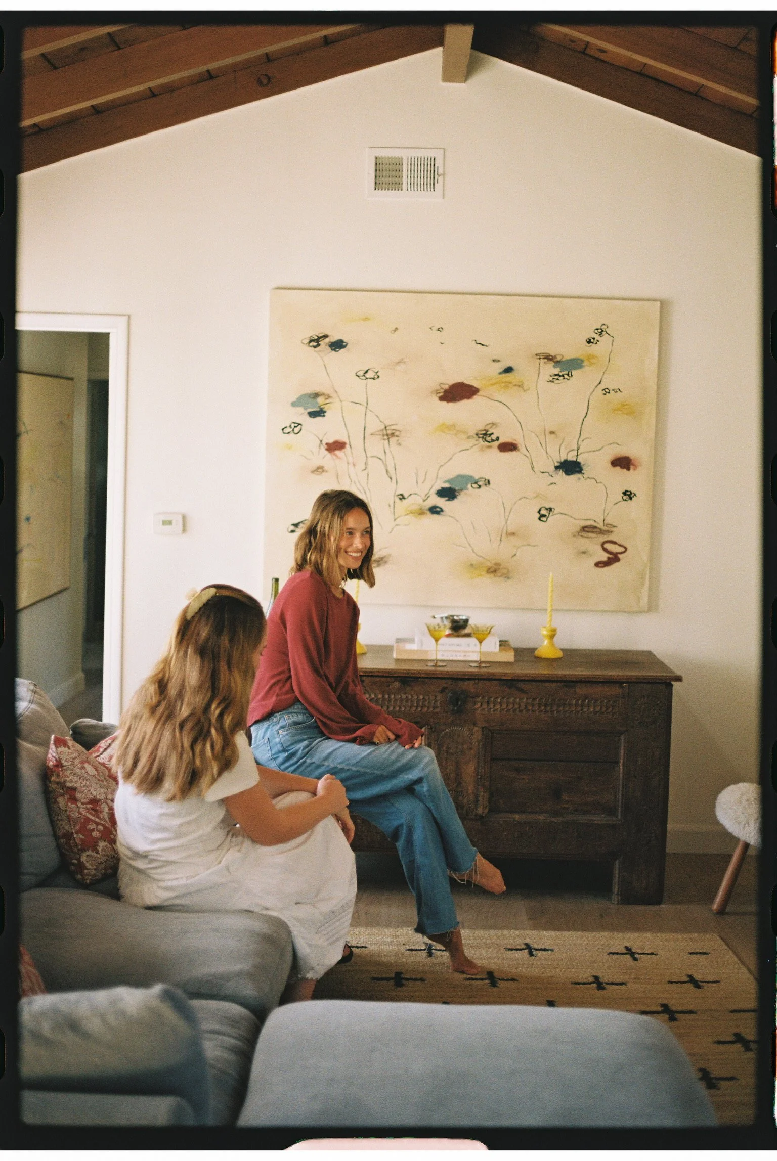 A woman with shoulder-length blonde hair, wearing a red long-sleeve shirt and blue jeans, sitting on a wooden sideboard in a living room, smiling while looking to her left. Two girls with long hair sit nearby, one wearing a white dress and the other with a floral hair accessory. The room features a modern abstract painting, a yellow candlestick, a small tray with drinks, a patterned rug, and a gray sofa with decorative pillows.