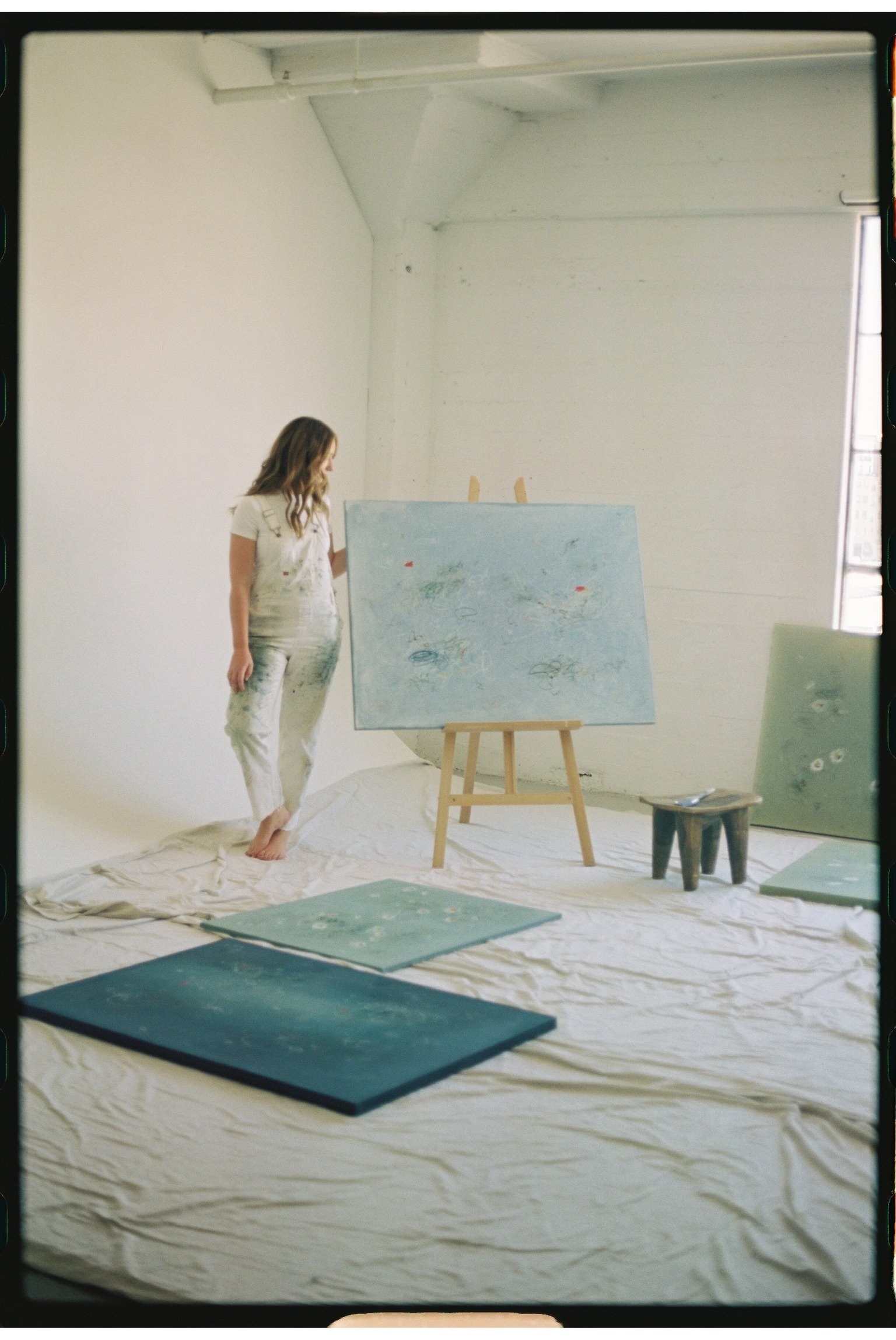 A woman in paint-splattered clothes stands barefoot in an art studio beside an easel with a canvas, surrounded by other canvases and art supplies on a crumpled white sheet on the floor.