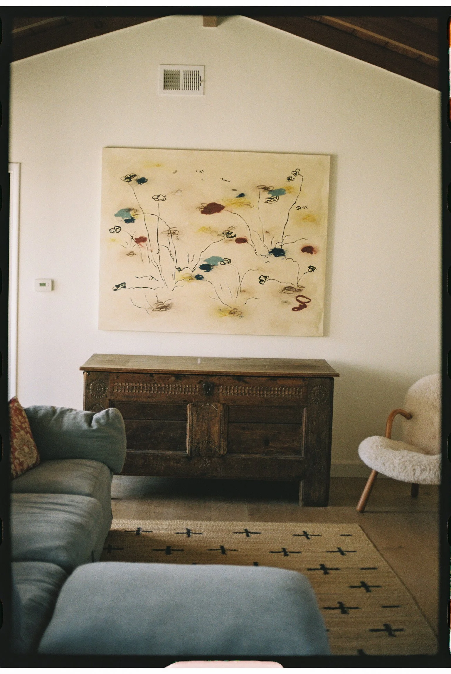 Living room with a beige couch on the left, a vintage wooden sideboard in the middle, a decorative beige armchair on the right, and a large abstract floral painting on the wall behind the sideboard. The room has a high ceiling with wooden beams and a vent.