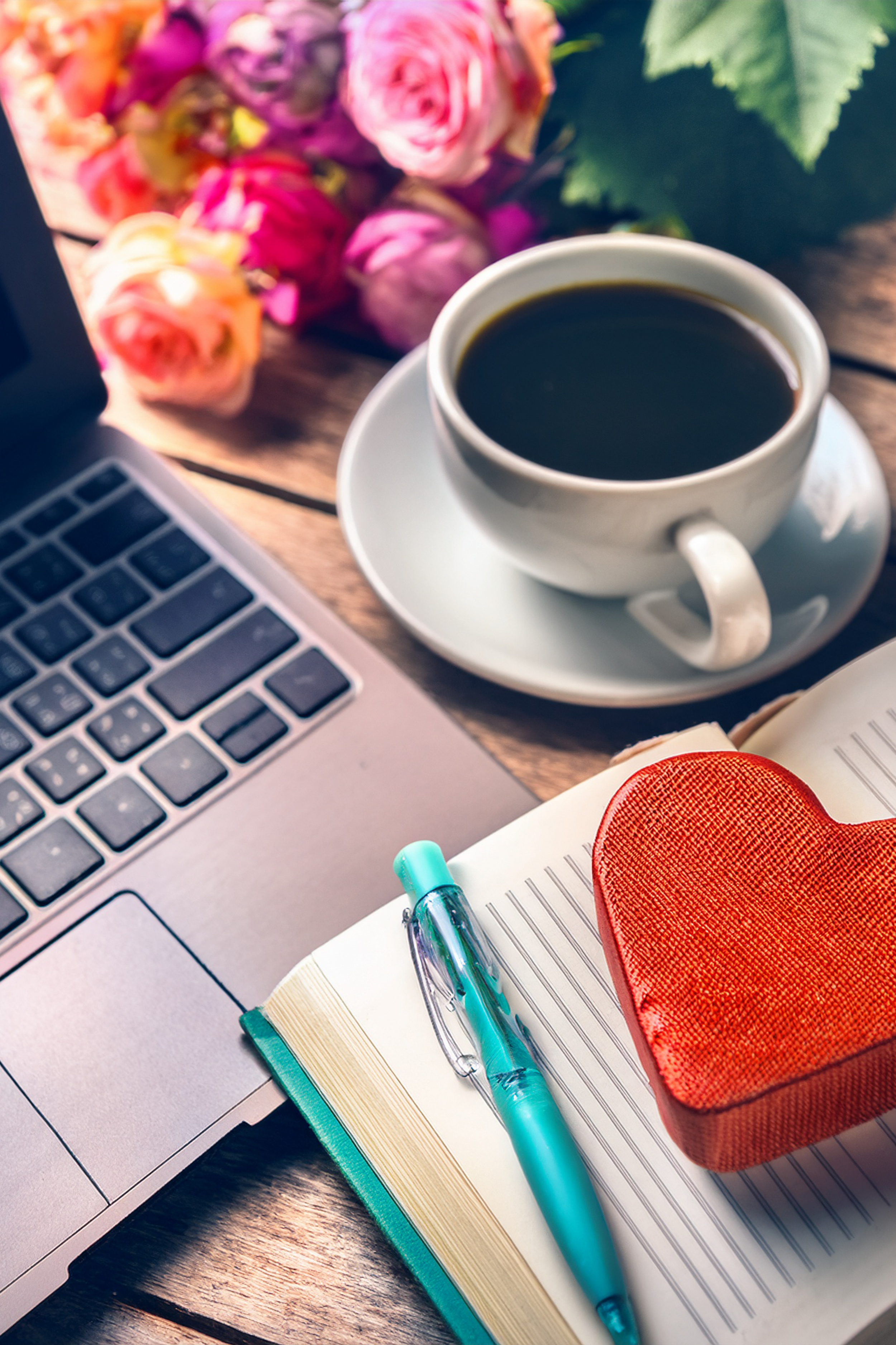 Tasse schwarzer Kaffee, pinke Rosen, Laptop, offene Notizbuch mit blauer Kugelschreiber, rotes Herz, Geschenkbox, Schreibblock, blauer Textmarker auf einem Holzschreibtisch.