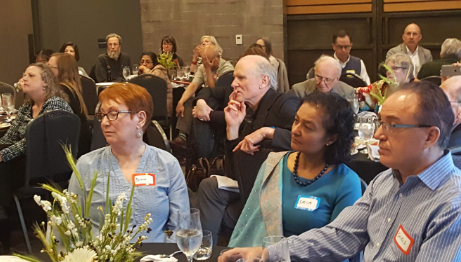 Audience seated at tables during a conference or event, listening attentively, with floral arrangements and glasses on the tables.