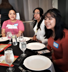 Three women sitting at a dinner table with empty plates and glasses of water, engaging in conversation and smiling.
